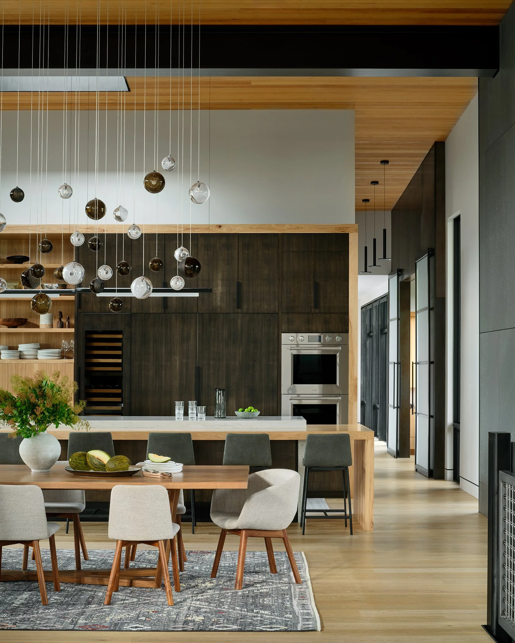 Modern kitchen and dining area with a large wooden dining table, upholstered chairs, a pendant light fixture with glass globes, and dark wood cabinetry.