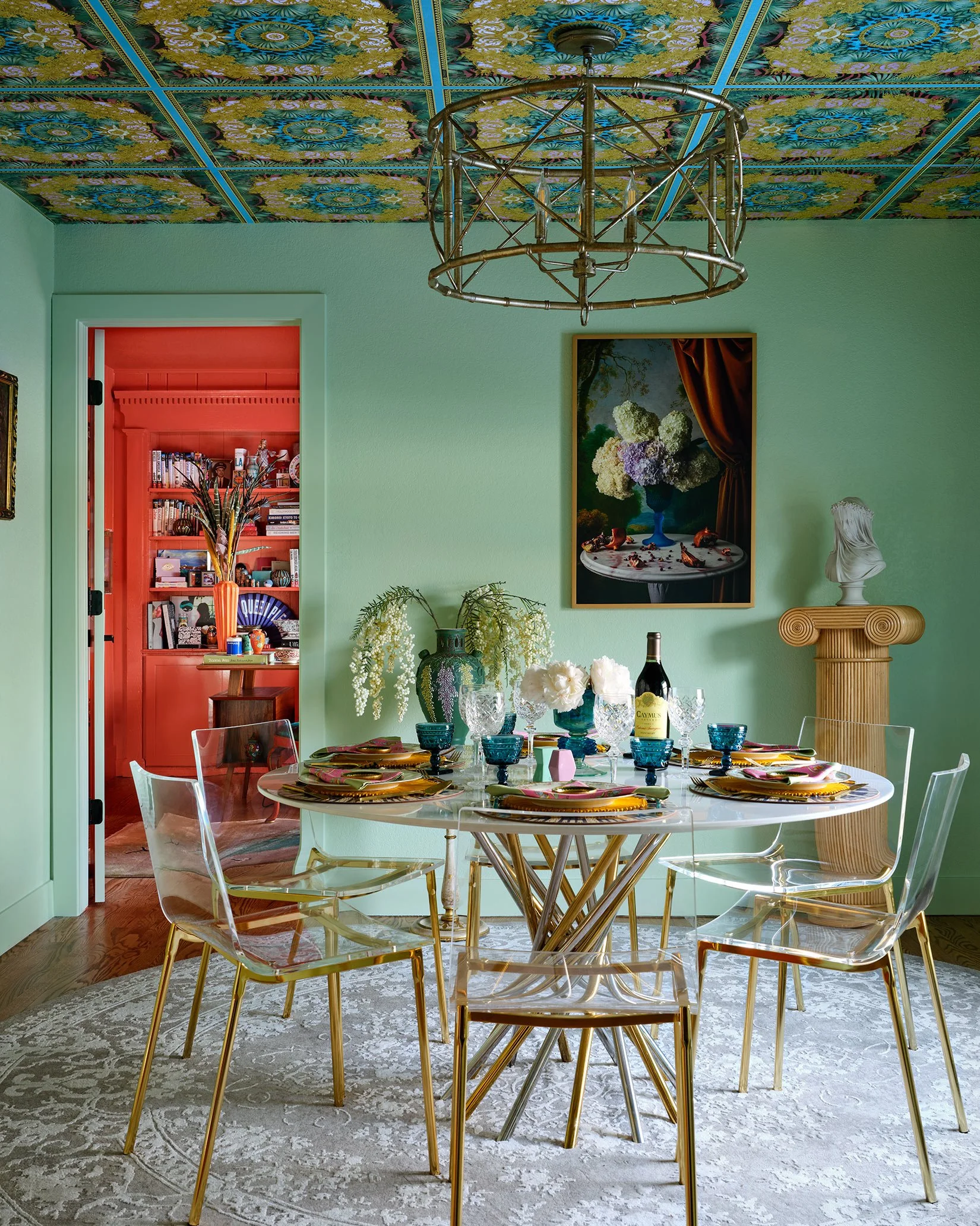 A colorful dining room with a round marble table set with vibrant dishes, glassware, and a wine bottle. Transparent acrylic chairs with gold legs surround the table. The room has light green walls, a patterned ornate ceiling with floral designs, and 