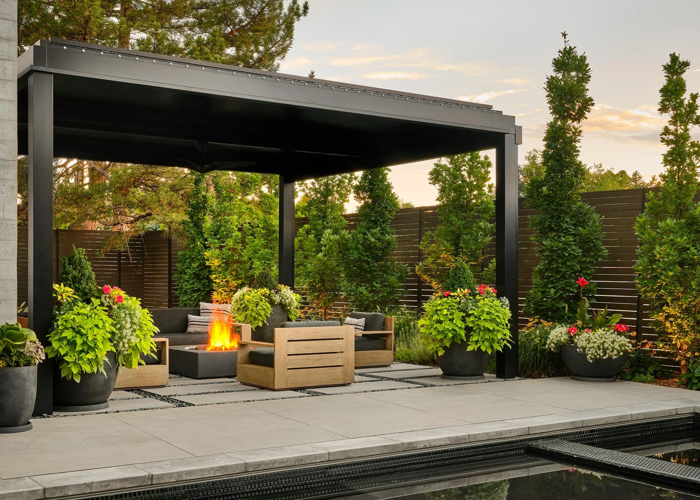Backyard patio with black pergola, outdoor seating, a fire pit, potted plants, and trees with a sunset sky.