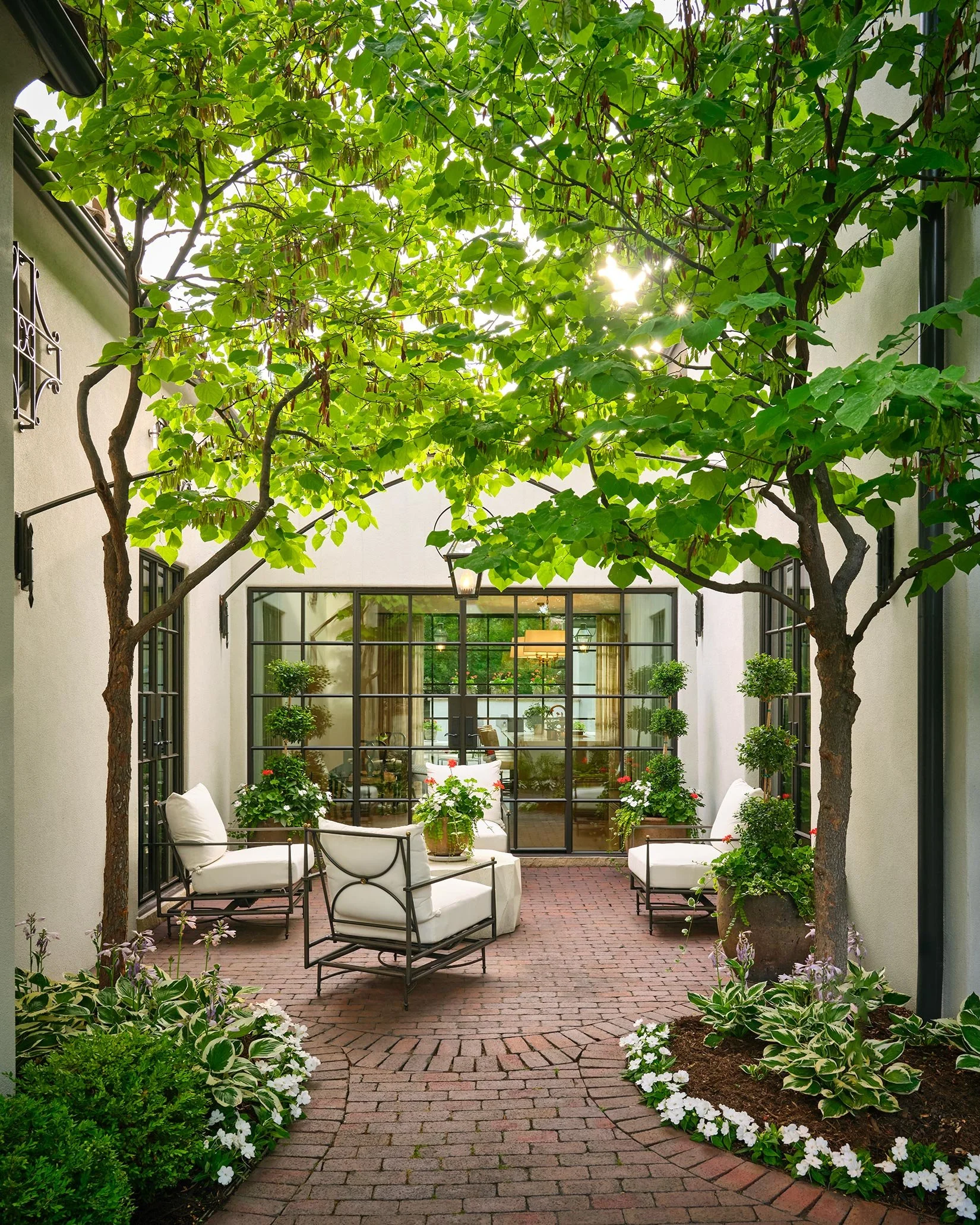 A cozy outdoor patio with white cushioned chairs, plants, trees, brick flooring, and a glass door leading into an indoor space.