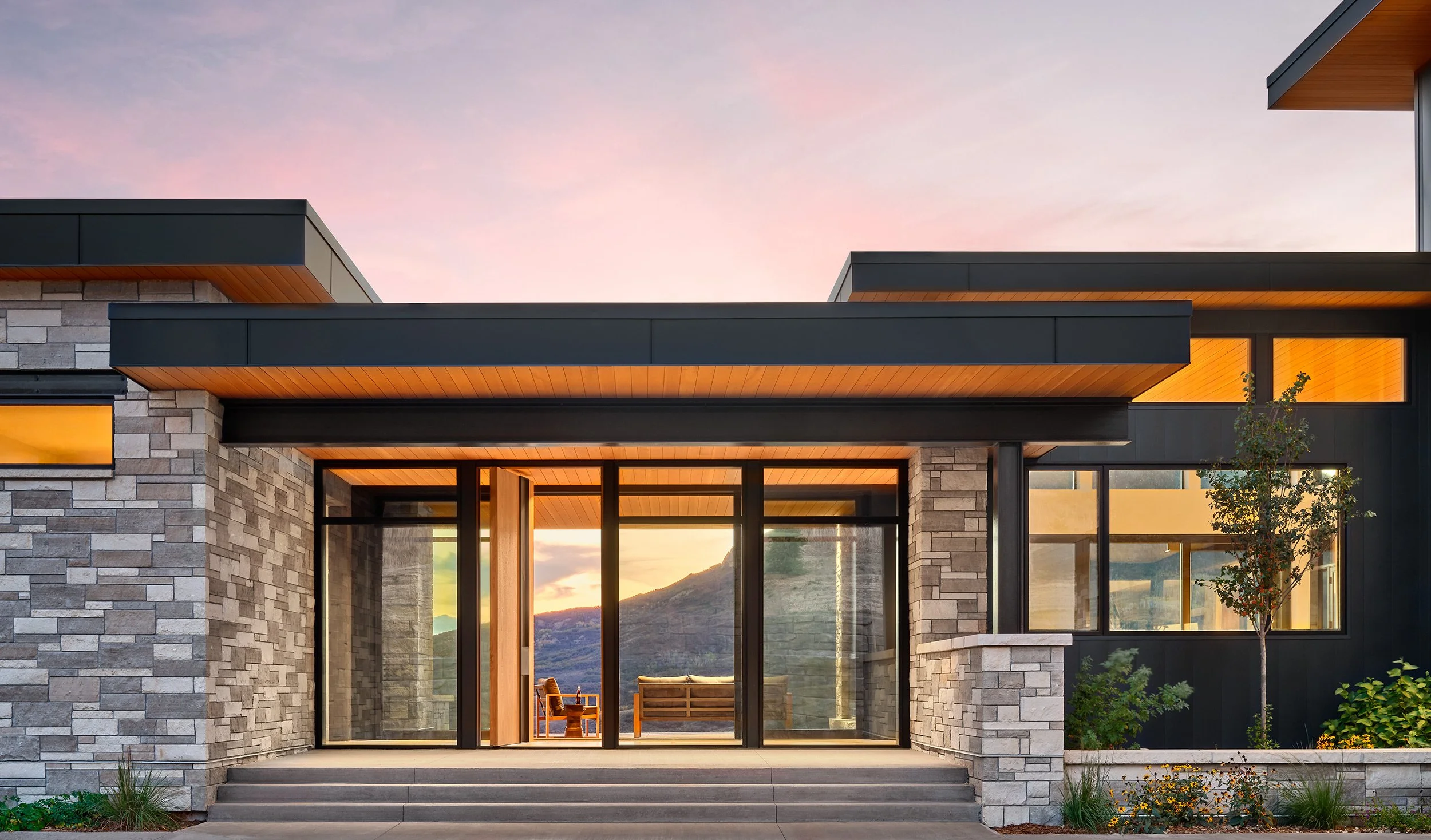 Modern house with stone and black siding, large glass doors, and a small front garden, during sunset.