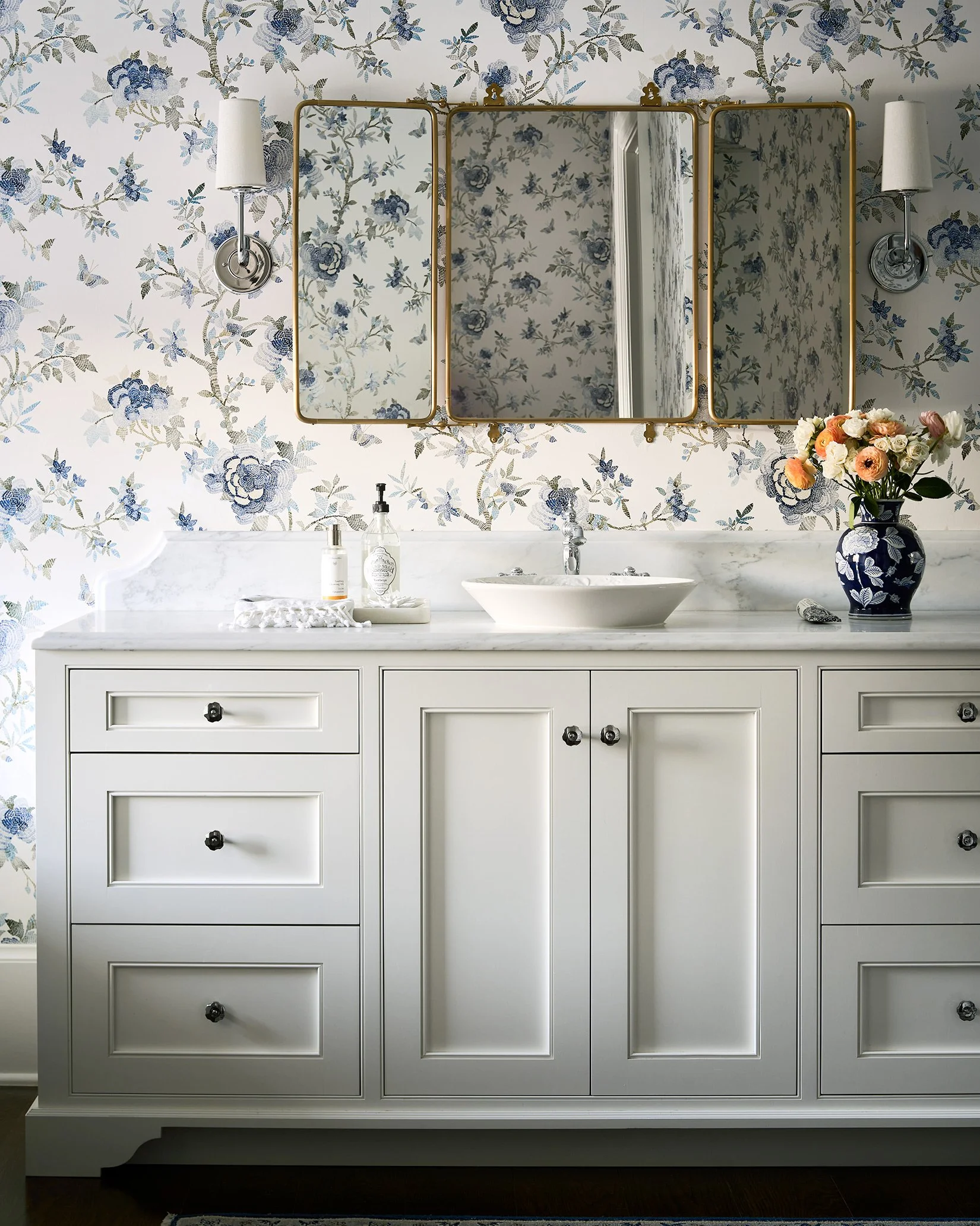 A bathroom vanity with a white wooden cabinet, marble countertop, and vessel sink, decorated with a blue floral wallpaper, a mirror, and a vase of flowers.