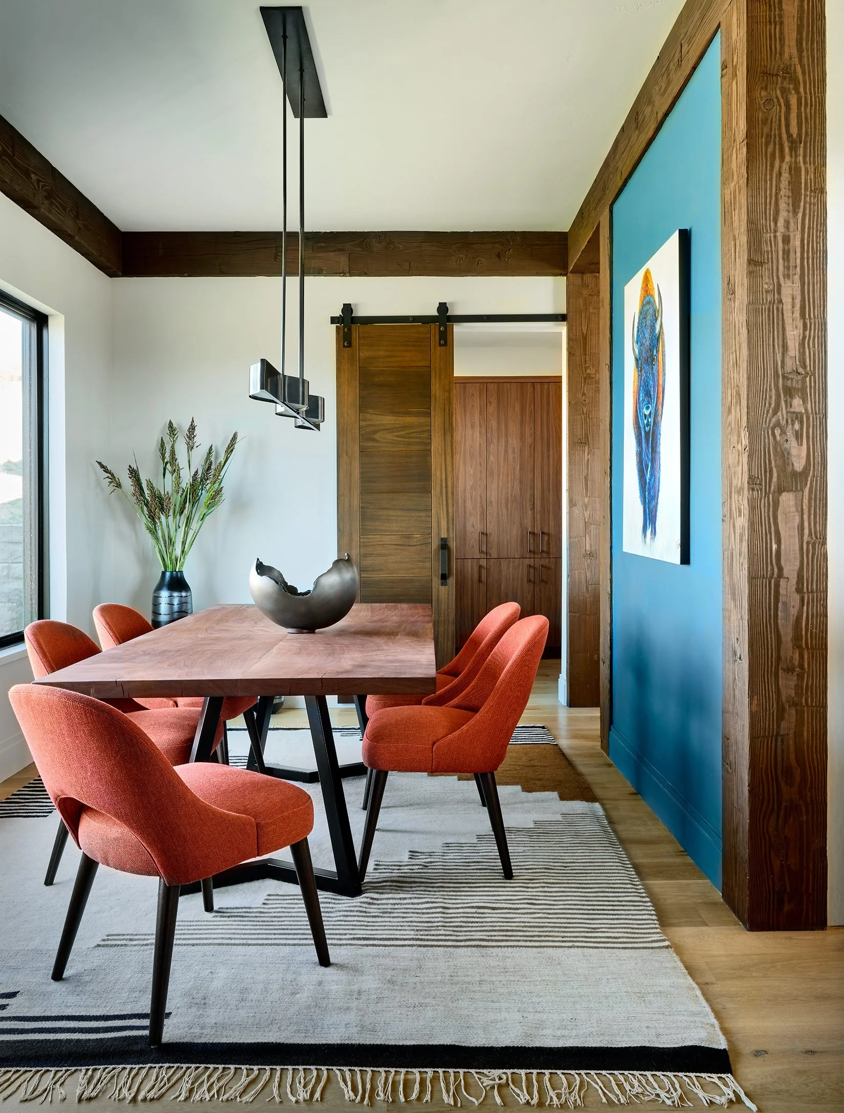 A modern dining room with a wooden table and red upholstered chairs, a large vase with plants, a picture of a bird on a blue wall, and a sliding barn door leading to a kitchen area.