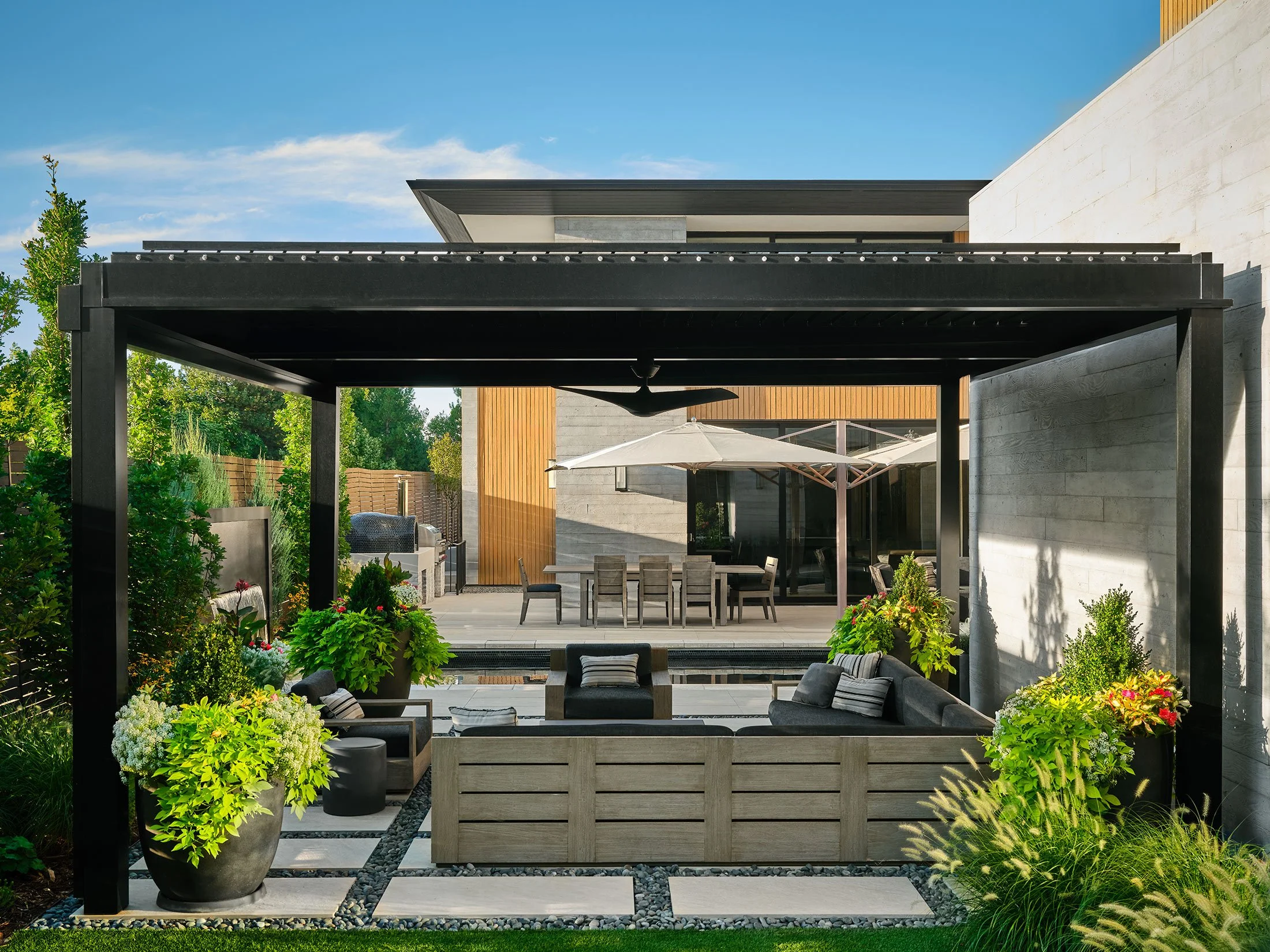 Modern outdoor patio with a black pergola, outdoor seating, large potted plants, a dining table with umbrellas, and greenery.