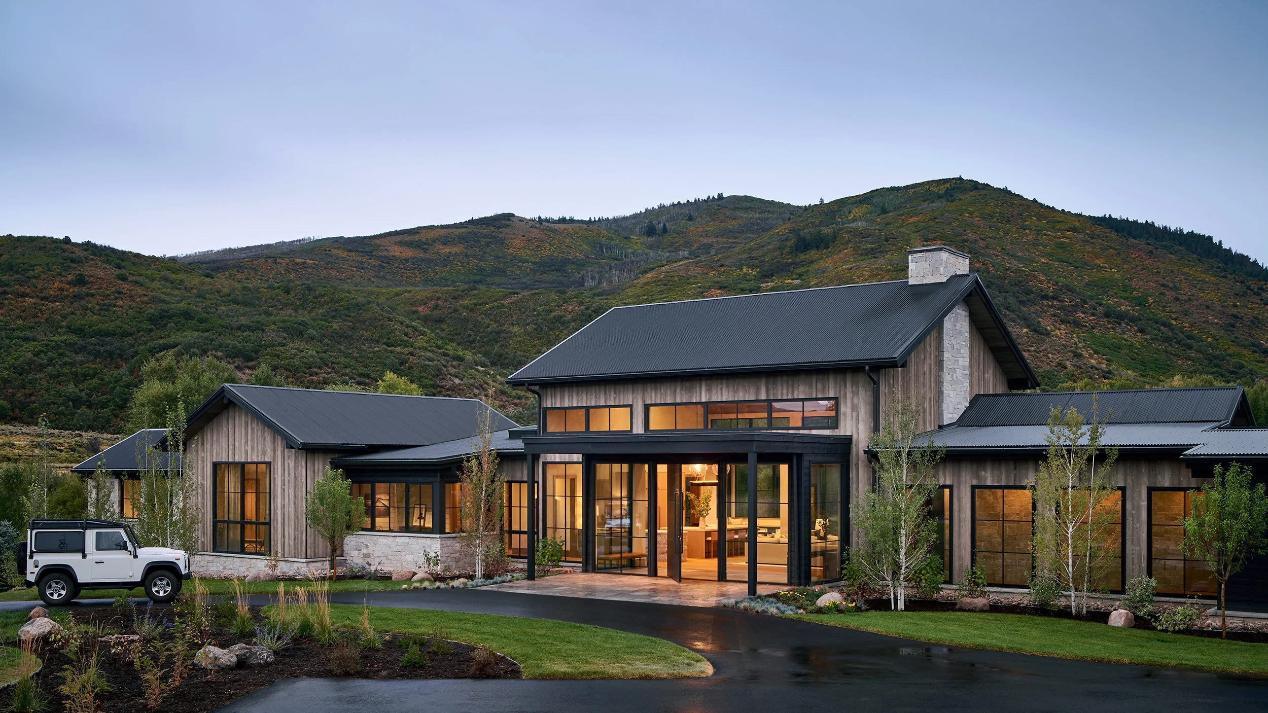 Modern house with large windows and a black metal roof, surrounded by greenery, with mountains in the background, and a white vehicle parked in the driveway.