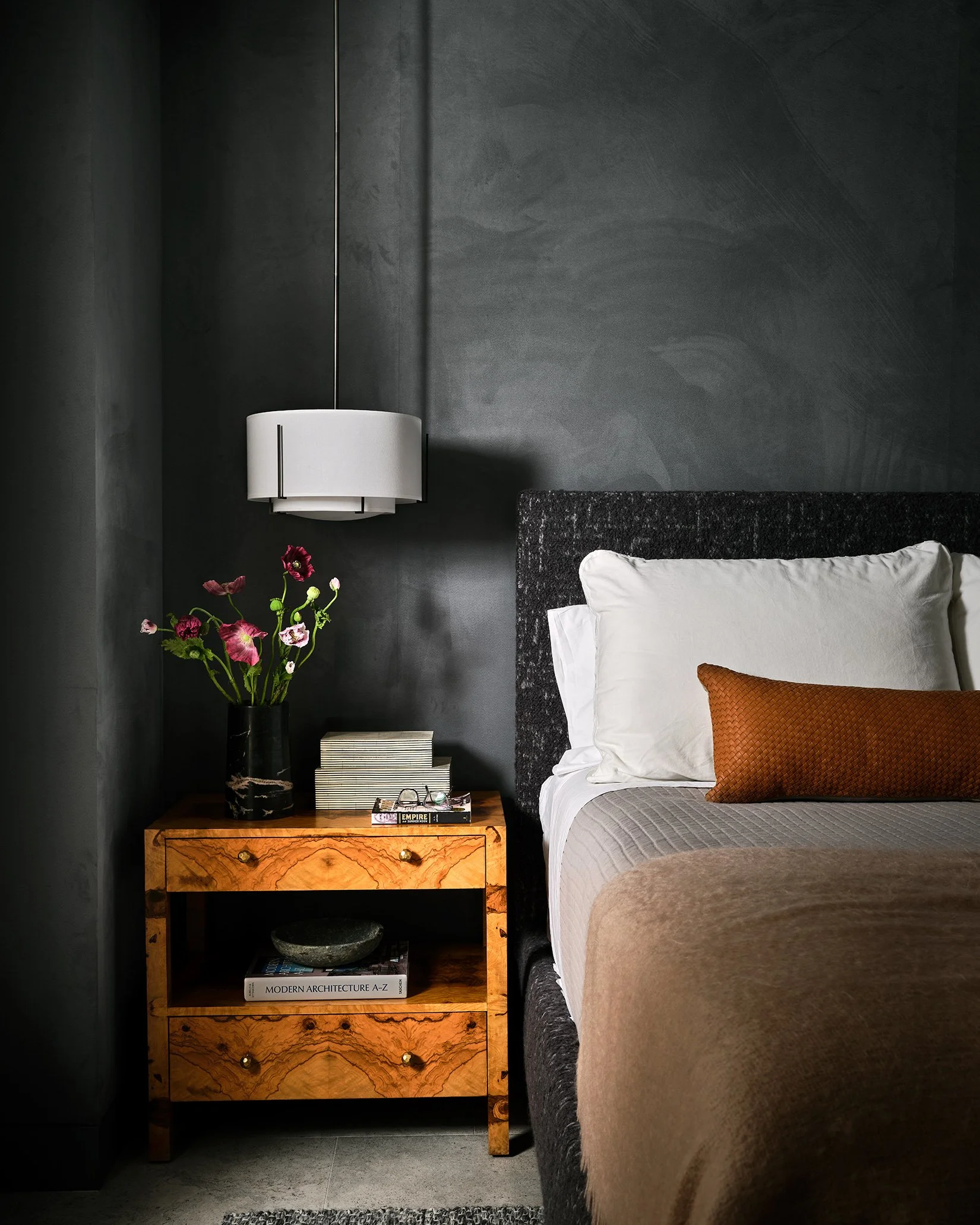Bedroom with dark gray walls, black upholstered headboard, white pillows, brown throw blanket, orange decorative pillow, wooden nightstand with a black vase holding pink and purple flowers, books, and a decorative bowl.