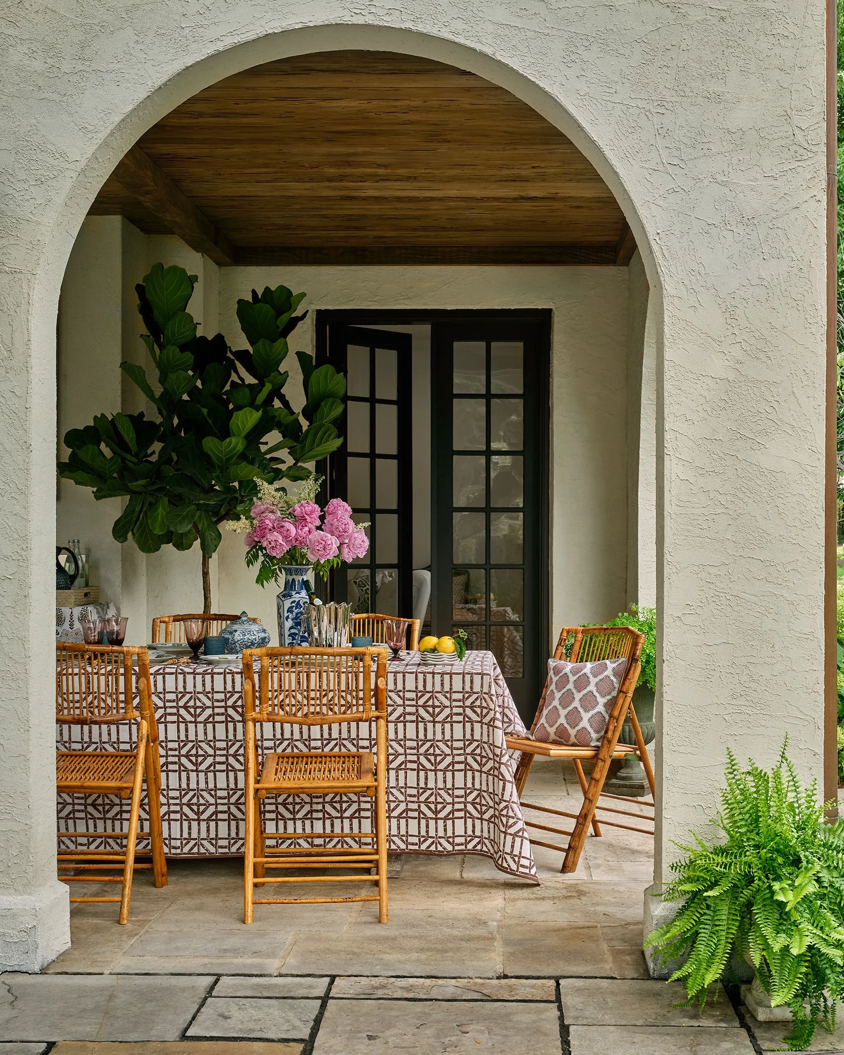 Outdoor dining area with a table covered by a patterned tablecloth, surrounded by bamboo chairs, decorated with a flower arrangement, potted plants, and located under an archway leading to a patio.