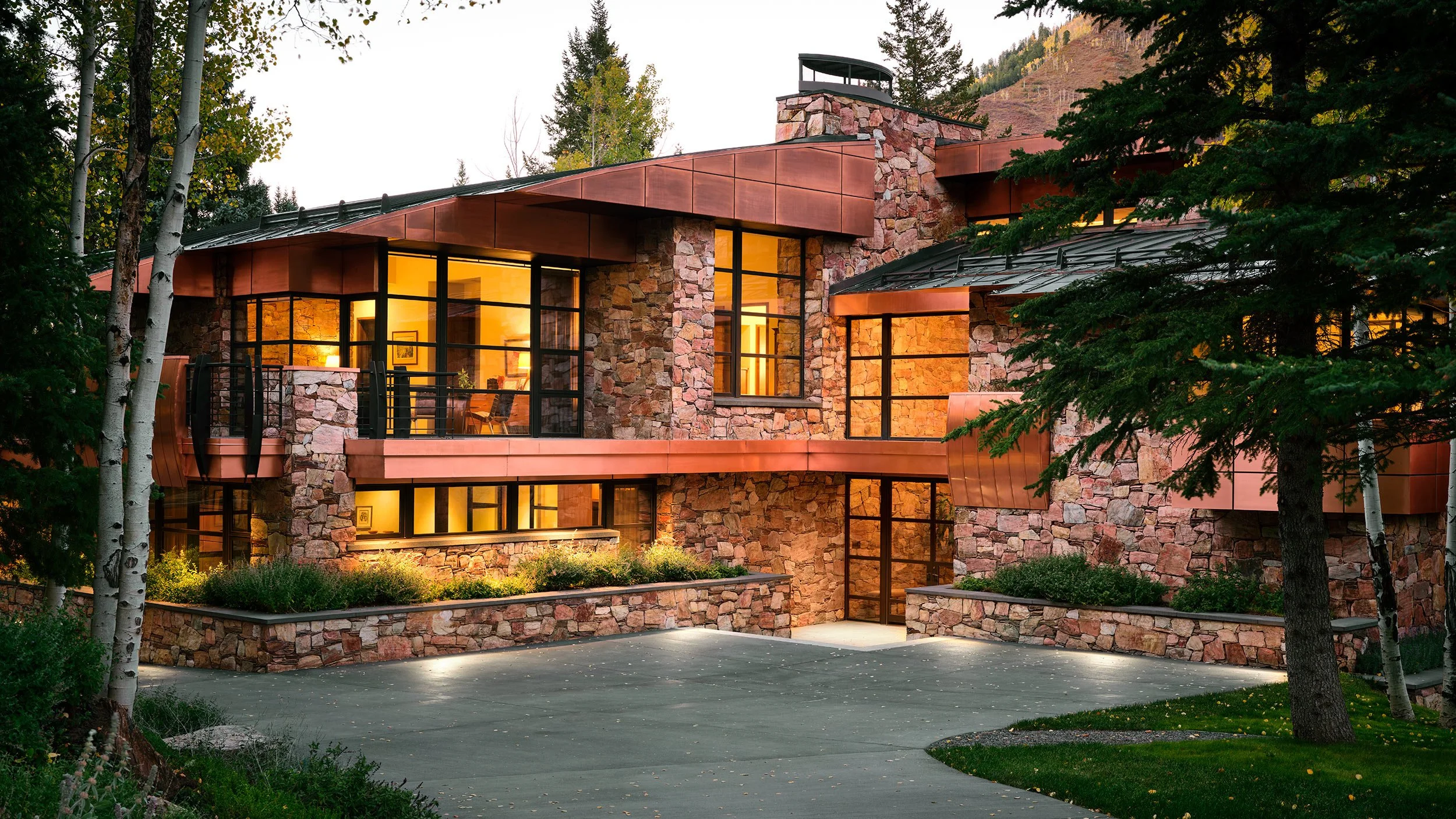 Modern house with stone and copper exterior, large glass windows, illuminated interior, surrounded by trees and landscaping at dusk.