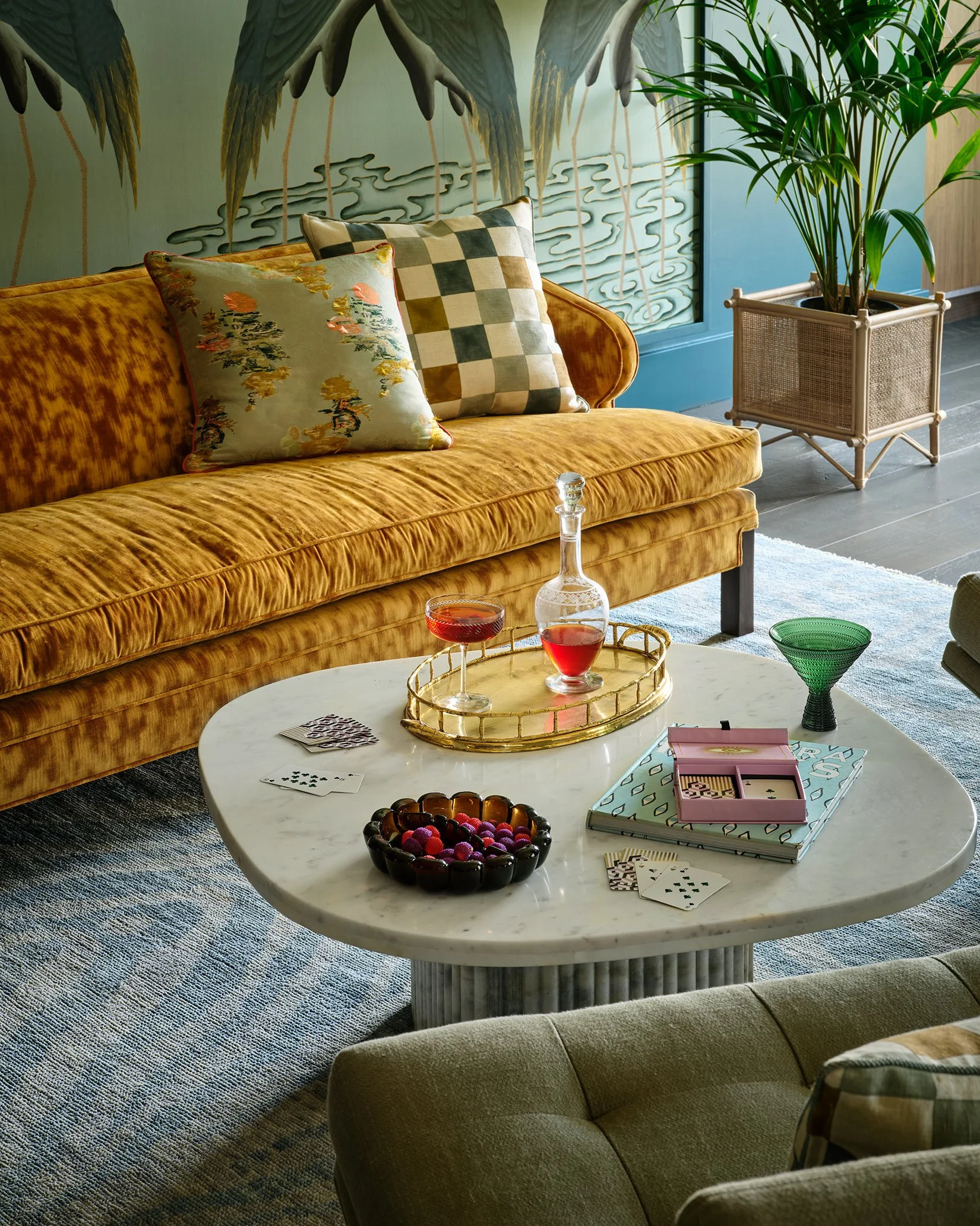 A living room with an orange velvet sofa, two decorative pillows, a white marble coffee table with drinks, snacks, and board game accessories, a potted plant, and a wall with palm tree artwork.