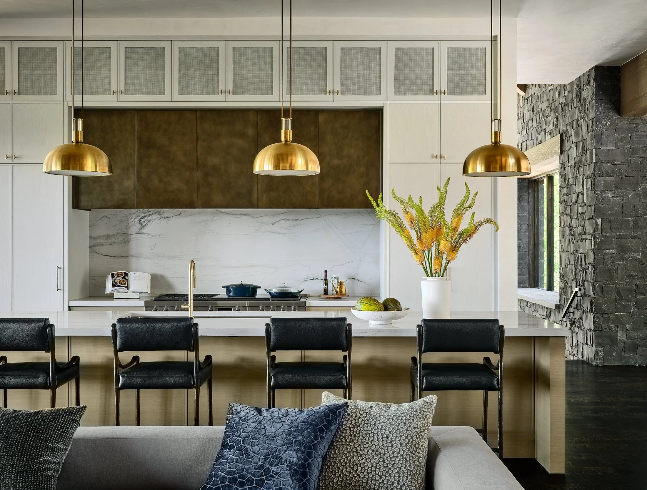 Modern kitchen with white cabinets, gold pendant lights, black chairs, and a white marble countertop with a vase of plants and some fruits.
