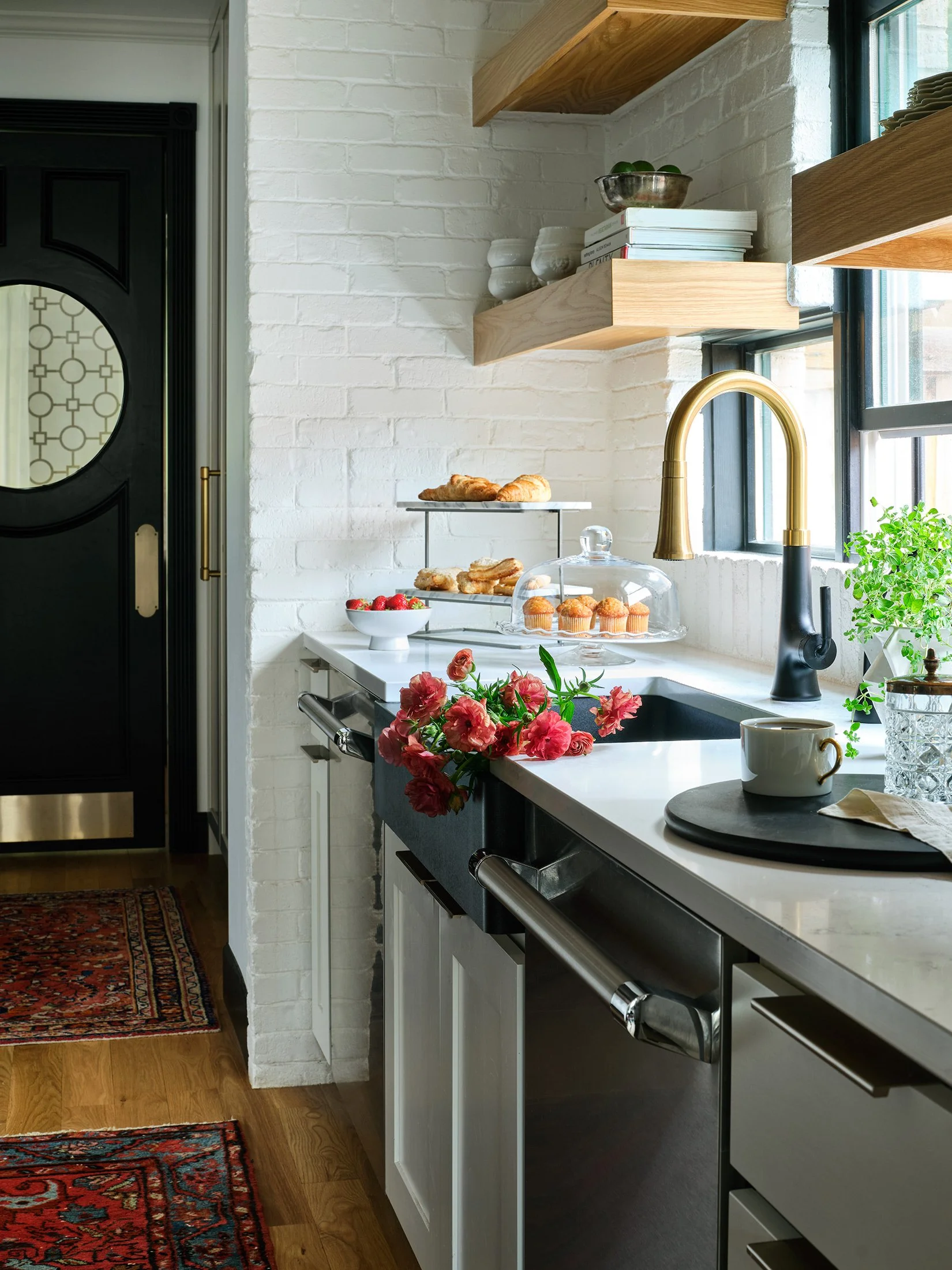 A modern kitchen with a white brick wall, wooden open shelves, and a window. On the counter, there is a vase with pink flowers, a cup, and a small plant. Behind the flowers, there are baked goods on a stand and a bowl of strawberries.