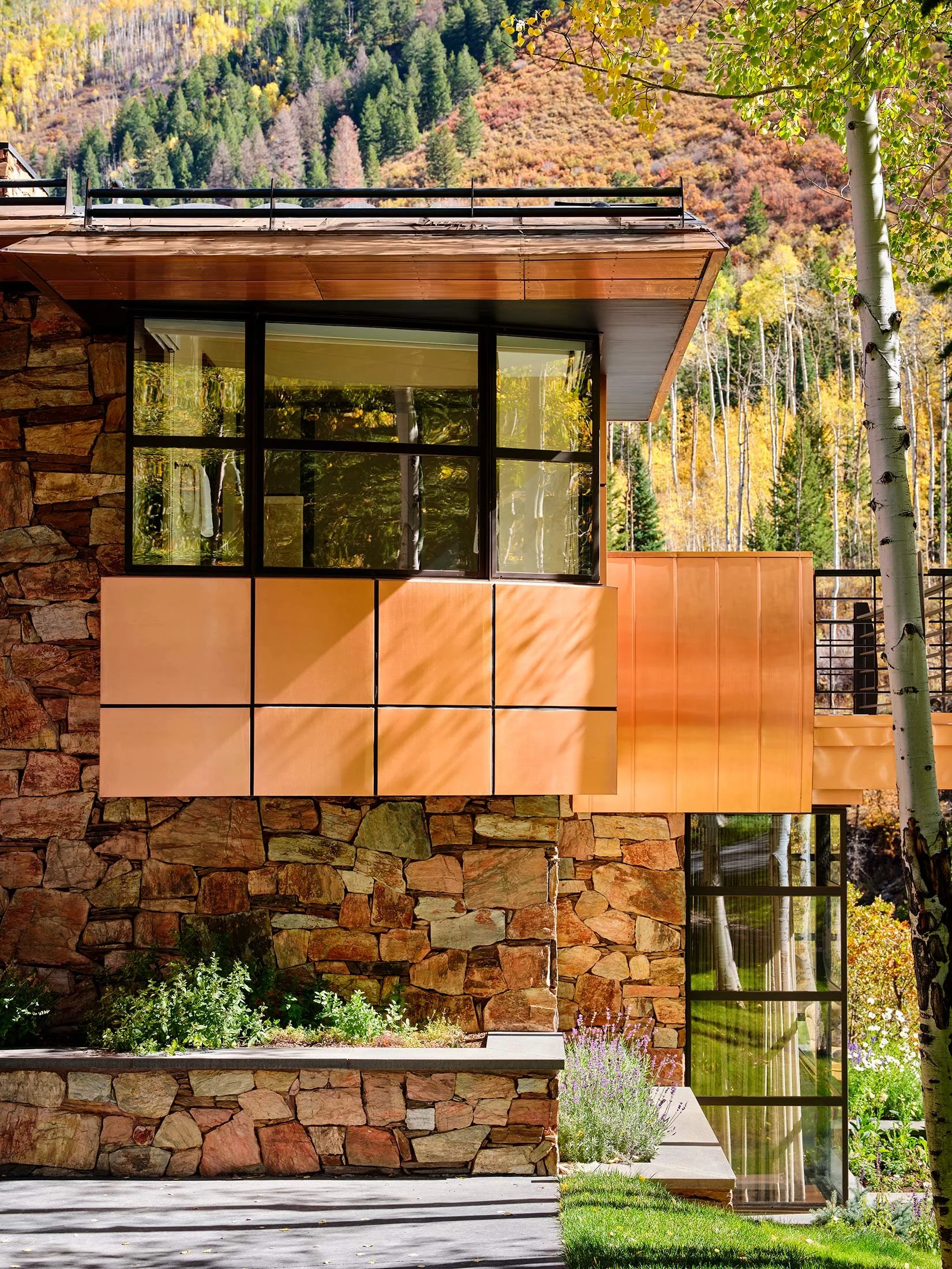 Modern house with stone and copper exterior, large glass windows, and surrounded by trees with fall foliage.