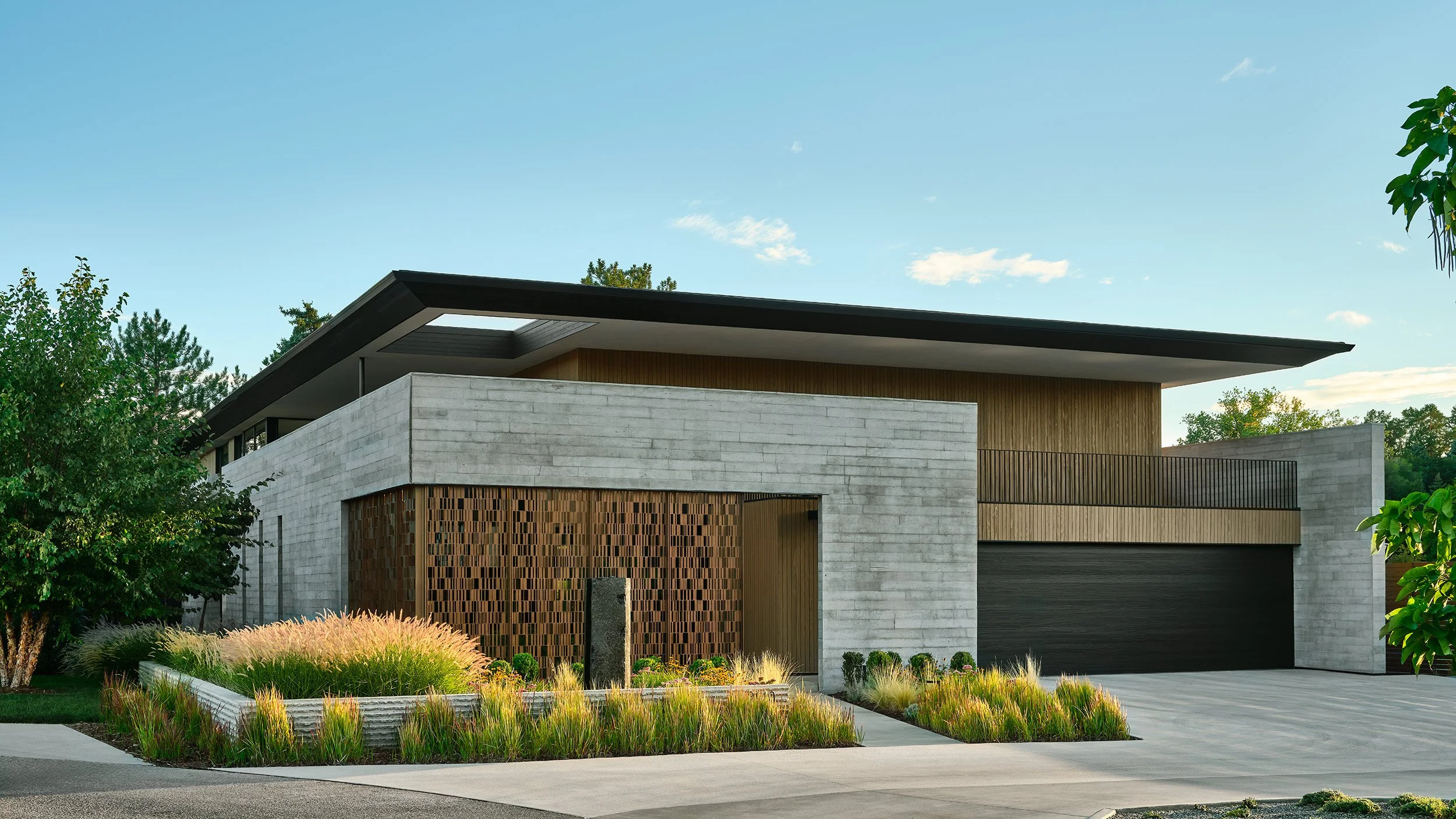 Modern house with flat roof, concrete and wooden exterior, large garage, landscaped front yard with grasses and trees.