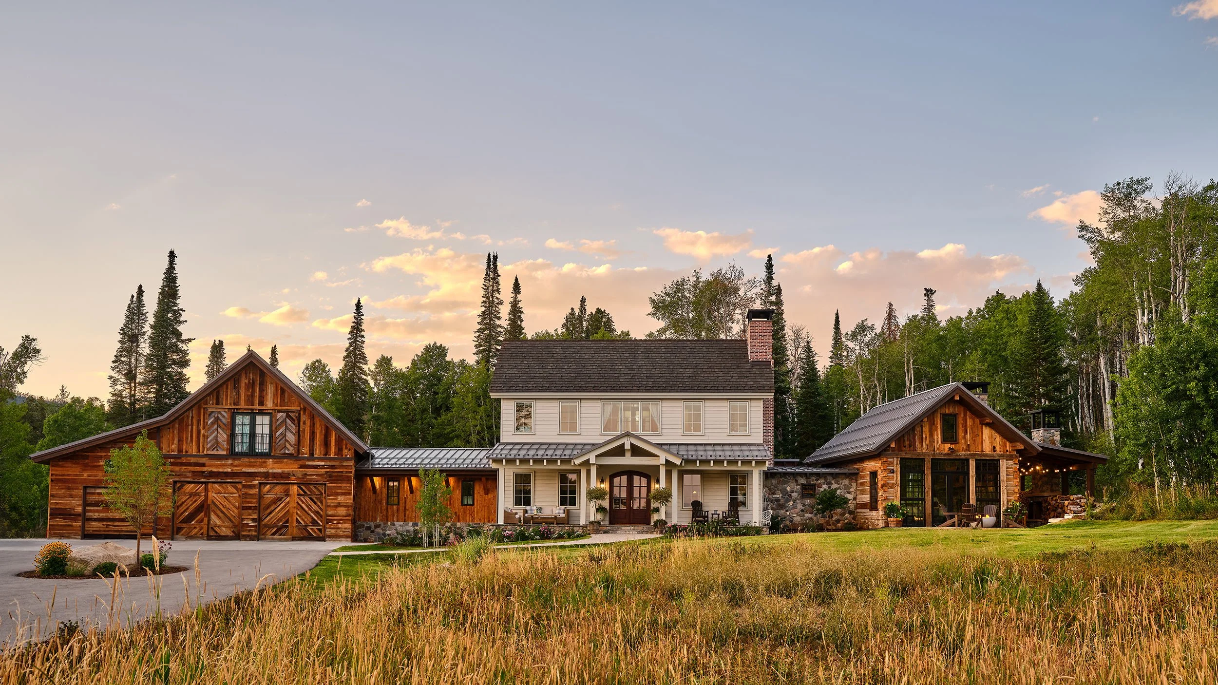 A large rustic house with two wings and a front porch, surrounded by trees and grass during sunset.