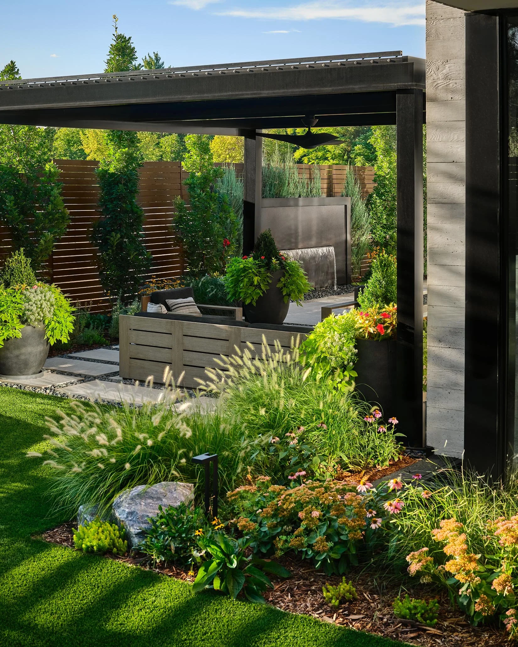 A modern backyard garden with lush greenery, colorful flowers, and a shaded seating area with outdoor furniture.
