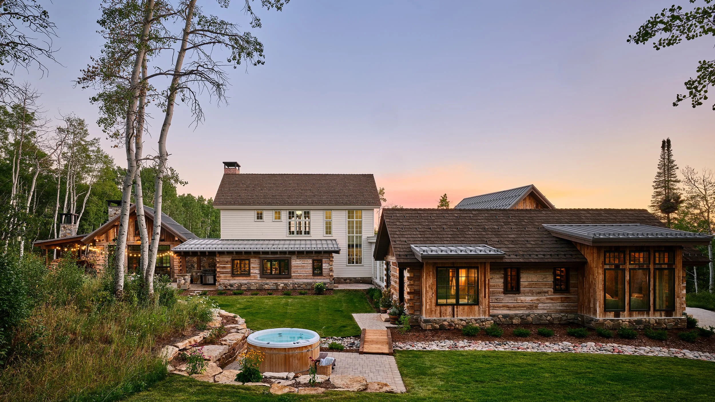 A large house with a mix of rustic stone and wood siding, large windows, a chimney, and a landscaped yard with a hot tub, in a wooded area at sunset.