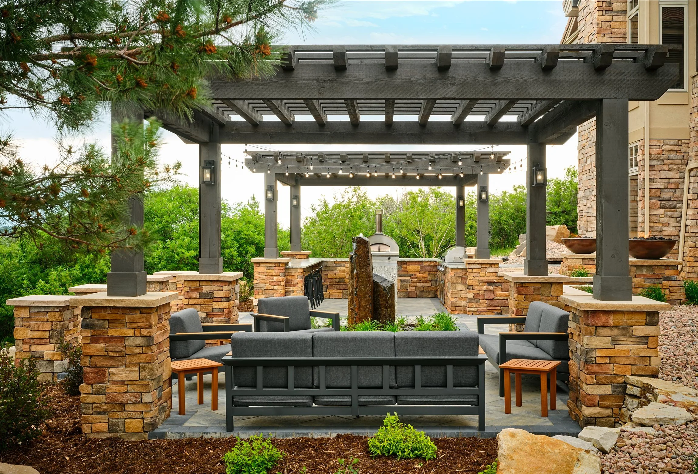 Outdoor living space with stone pillars, black pergolas, gray seating, a stone fireplace, and lush green trees in the background.