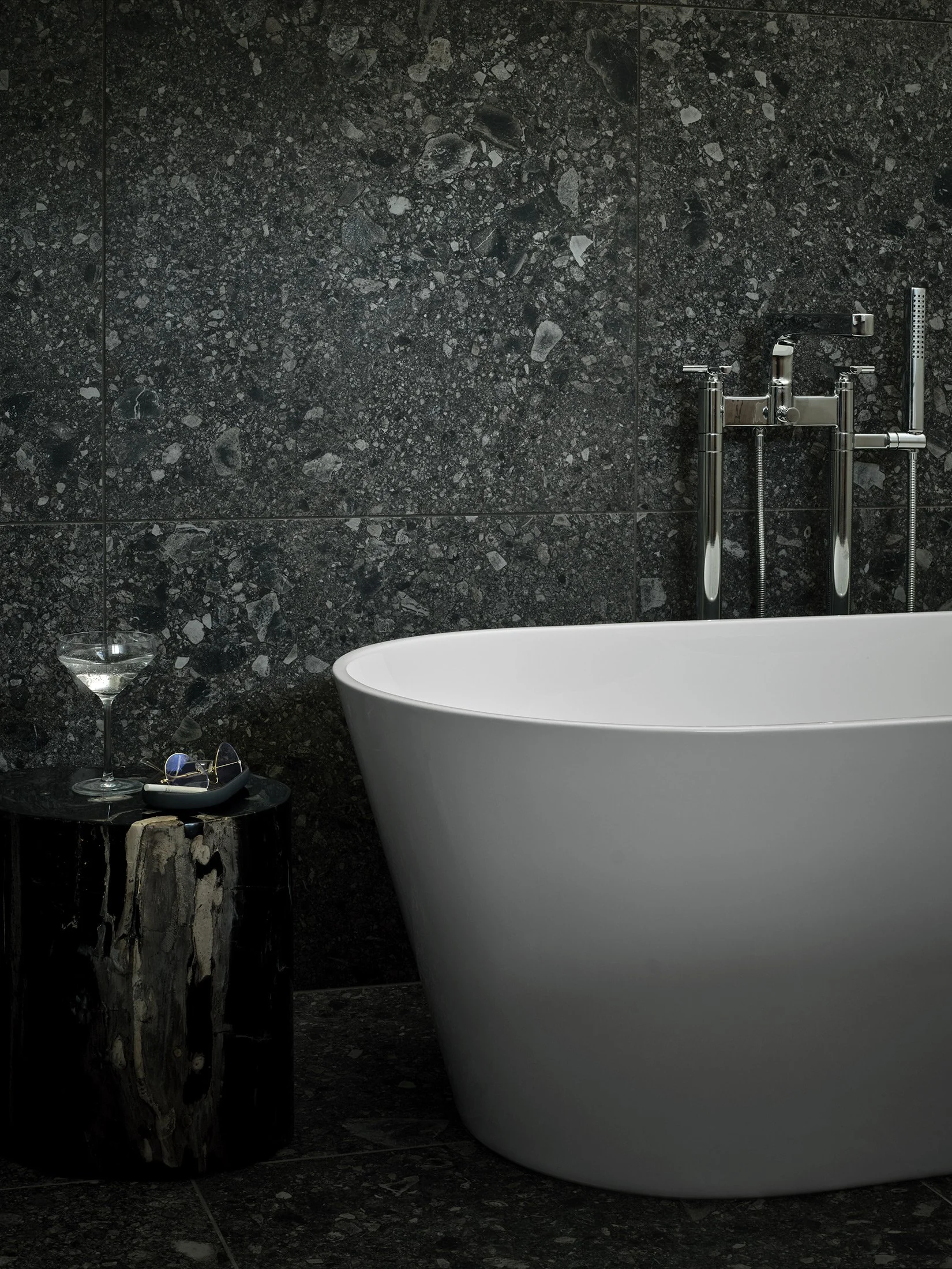 A modern black marble bathtub room with a white bathtub, a black marble side table holding a martini glass, sunglasses, and a remote control, and a textured black marble wall with chrome fixtures.