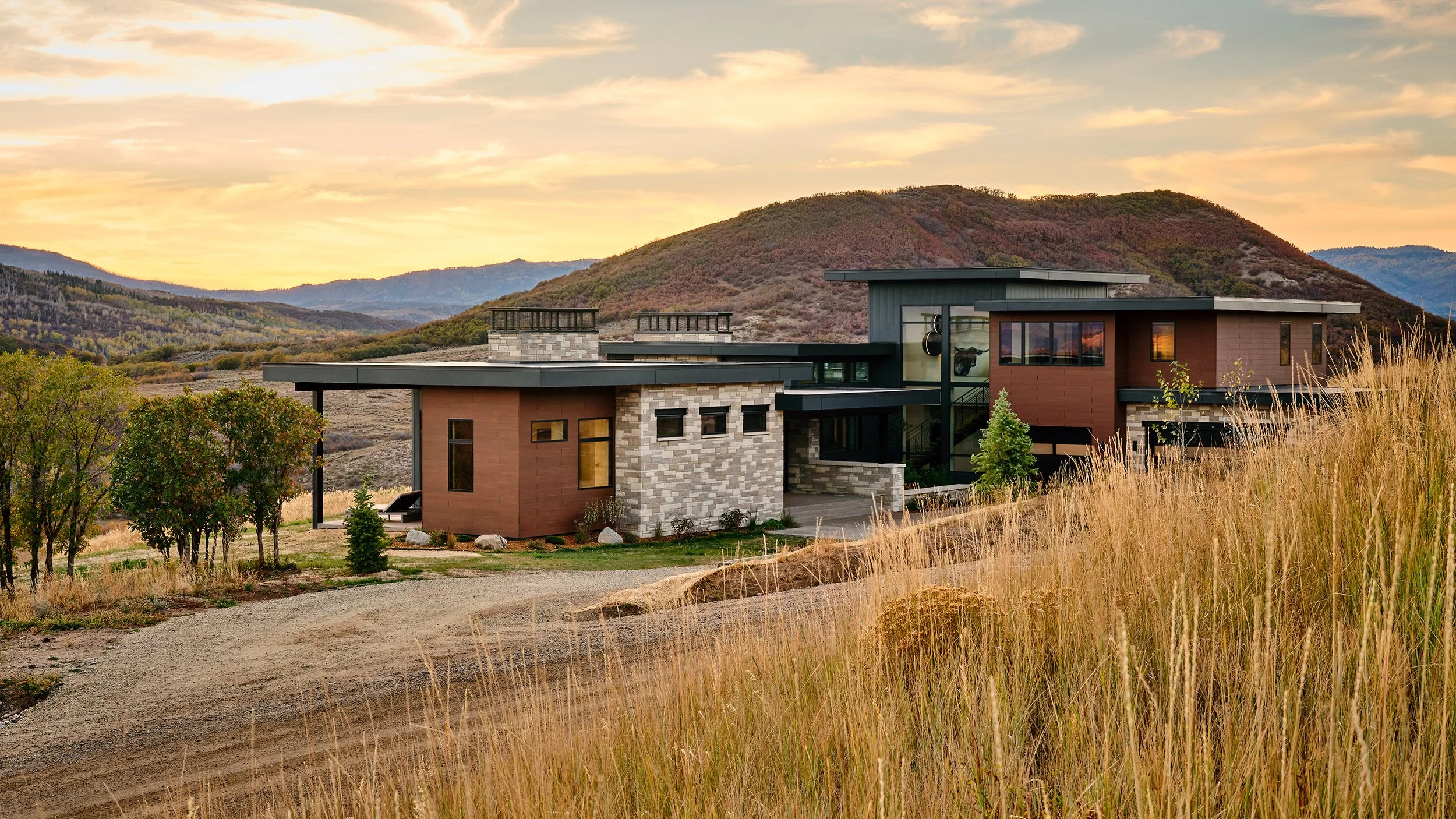 Modern house with a flat roof, large windows, and a mix of stone and wood exterior, situated in a hilly landscape during sunset