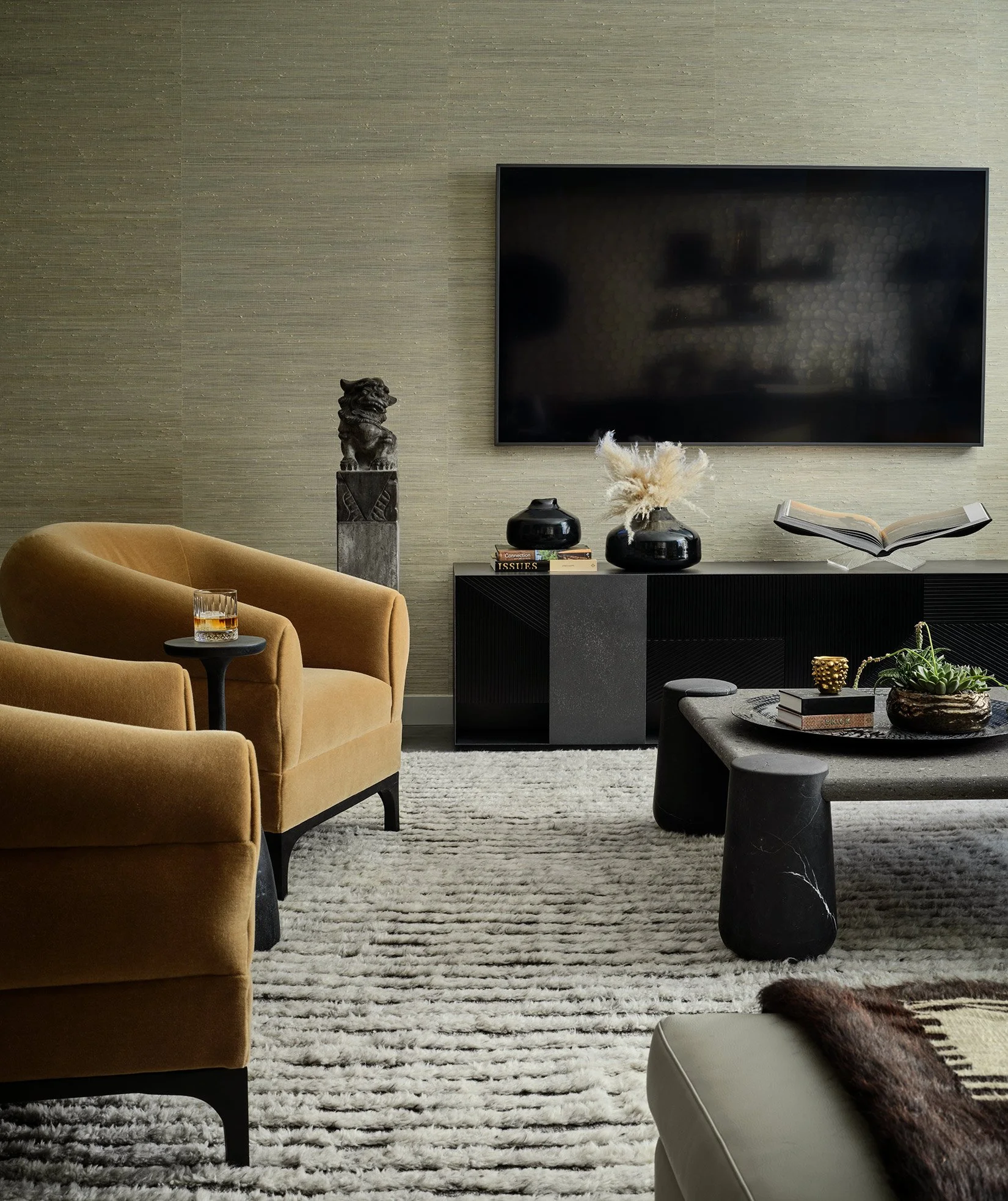 A modern living room with a beige armchair, a black sideboard with decorative items, and a large flat-screen TV mounted on the wall. There is a small black side table with a glass of whiskey, and a gray ottoman with a tray of plants and books. The ro