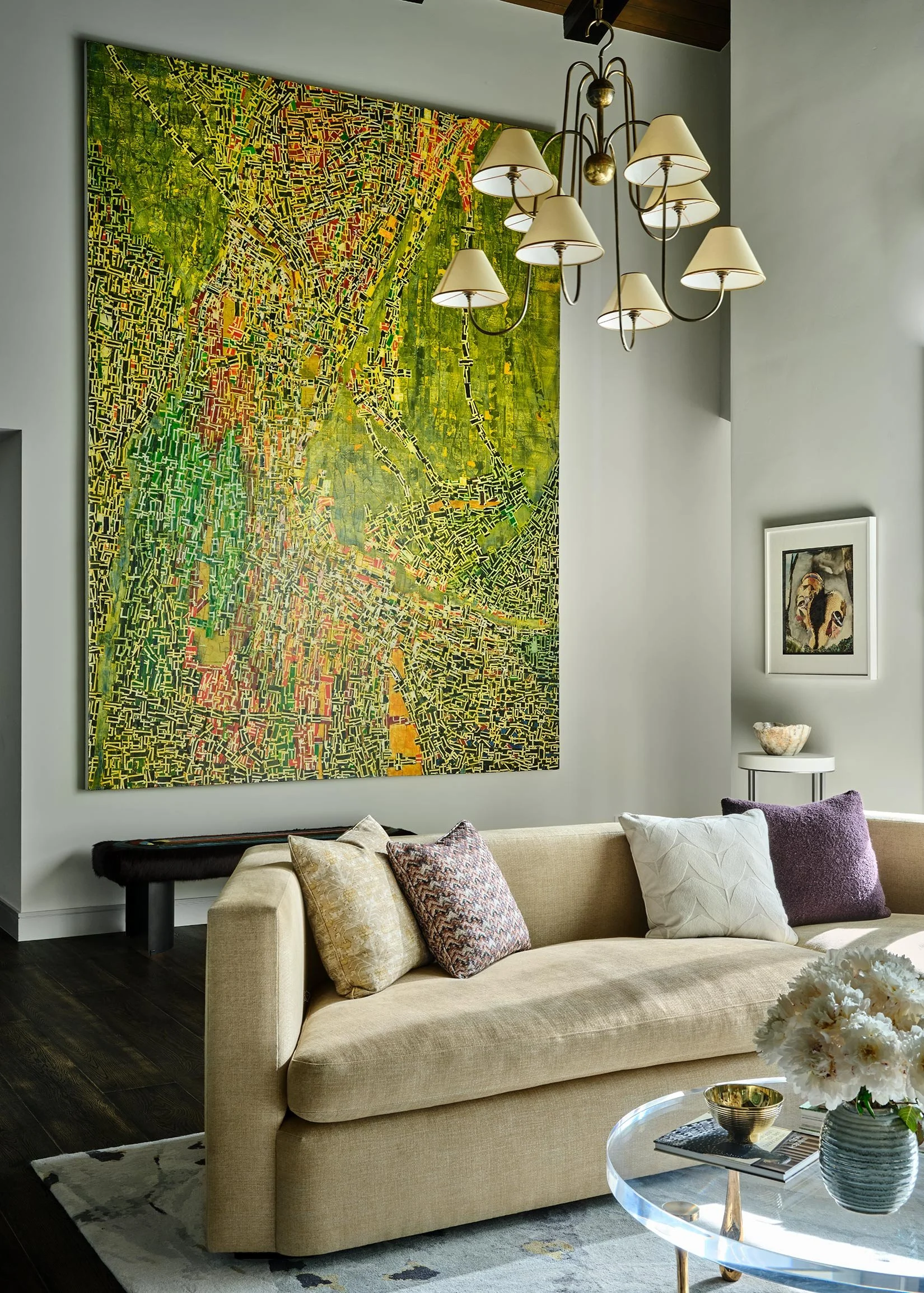 Living room with large colorful abstract painting, chandelier, beige sofa with pillows, small side table with decorative bowl, framed picture on the wall, and glass-topped coffee table with flowers.