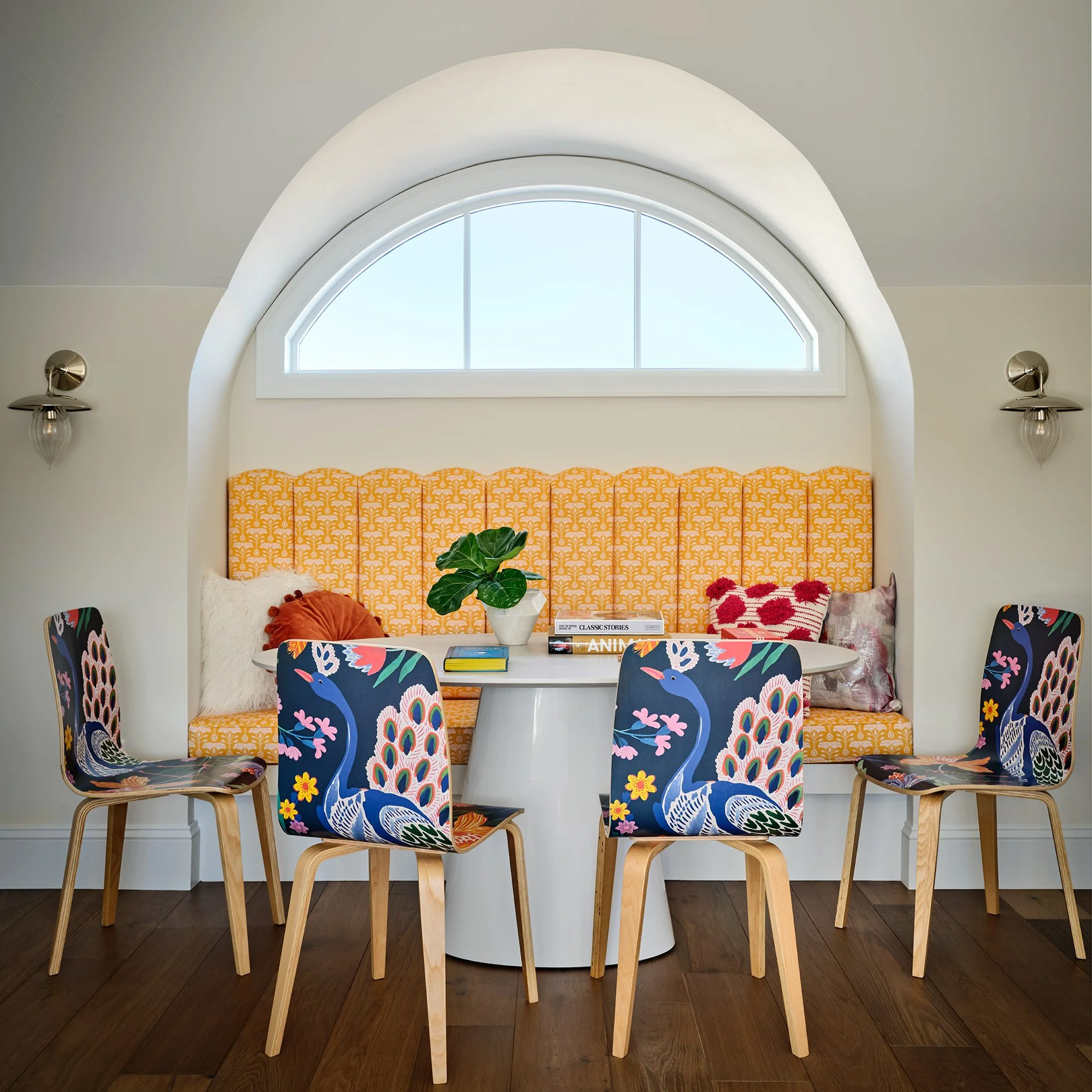 Dining area with peacock-patterned chairs, a round white table, and a built-in yellow patterned bench with decorative pillows, in front of a large arched window.