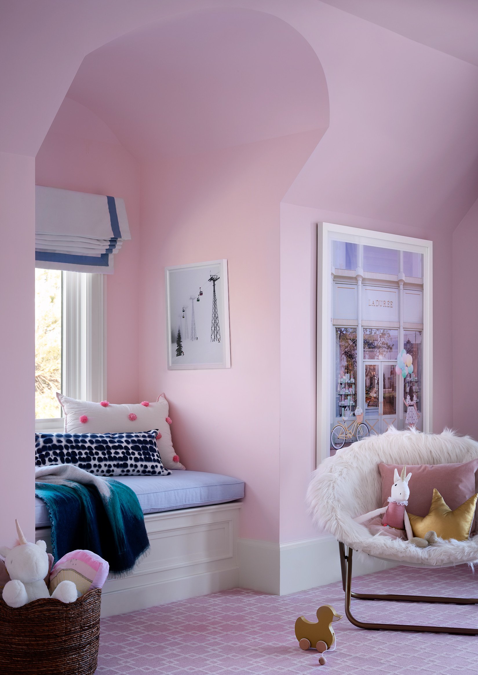 A cozy, pink-themed children's room with a window seat with pillows, a fluffy accent chair with pillows and a plush toy, a wicker basket filled with stuffed animals, and a small wooden duck toy on the pink patterned carpet. There is a window and a do