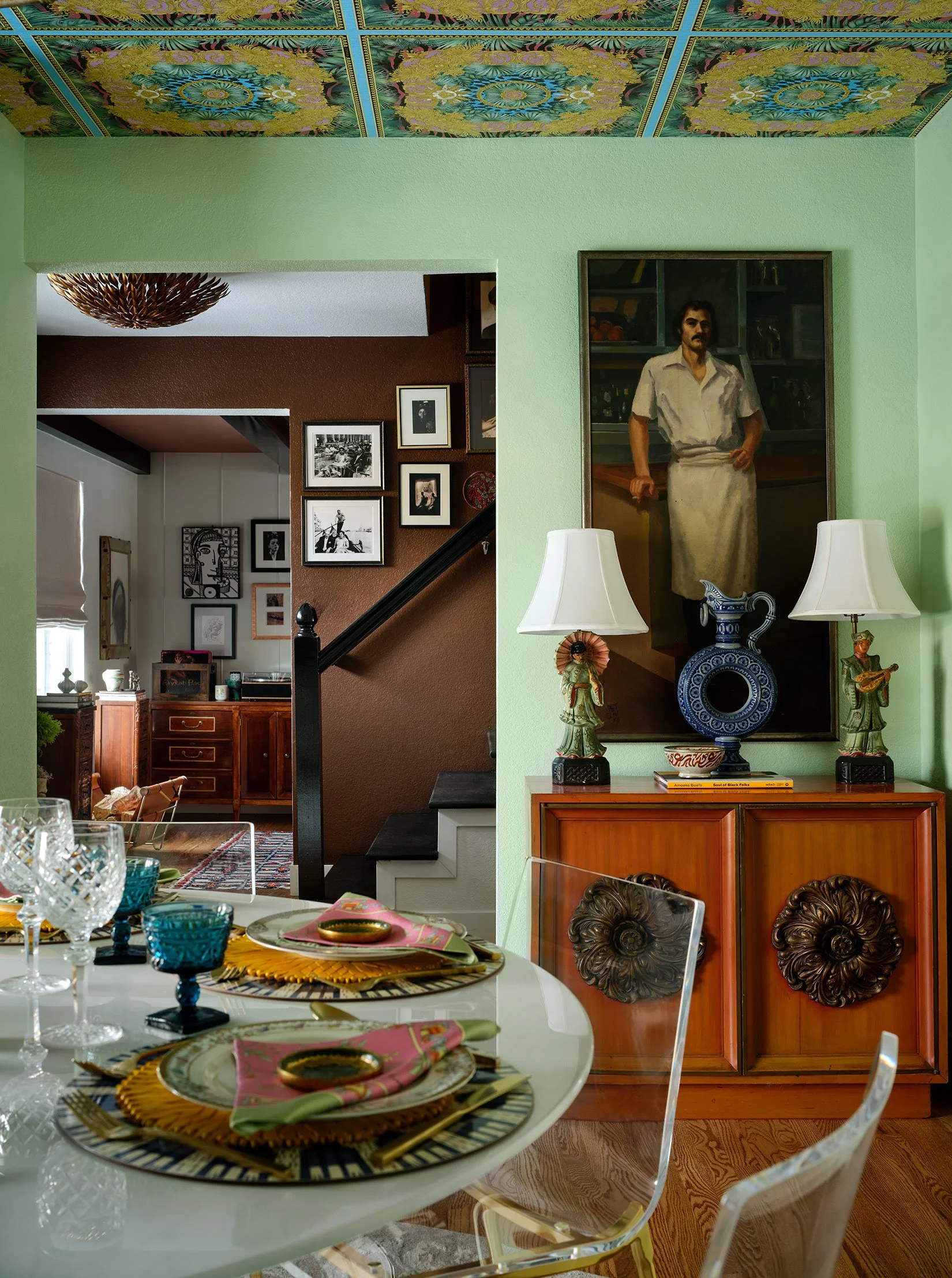 A dining room with a table set with colorful dishes, glasses, and napkins, and a sideboard with decorative statues, a large blue ceramic vase, and lamps. The background shows a wall with a large portrait of a man in a white shirt and apron, and a gal