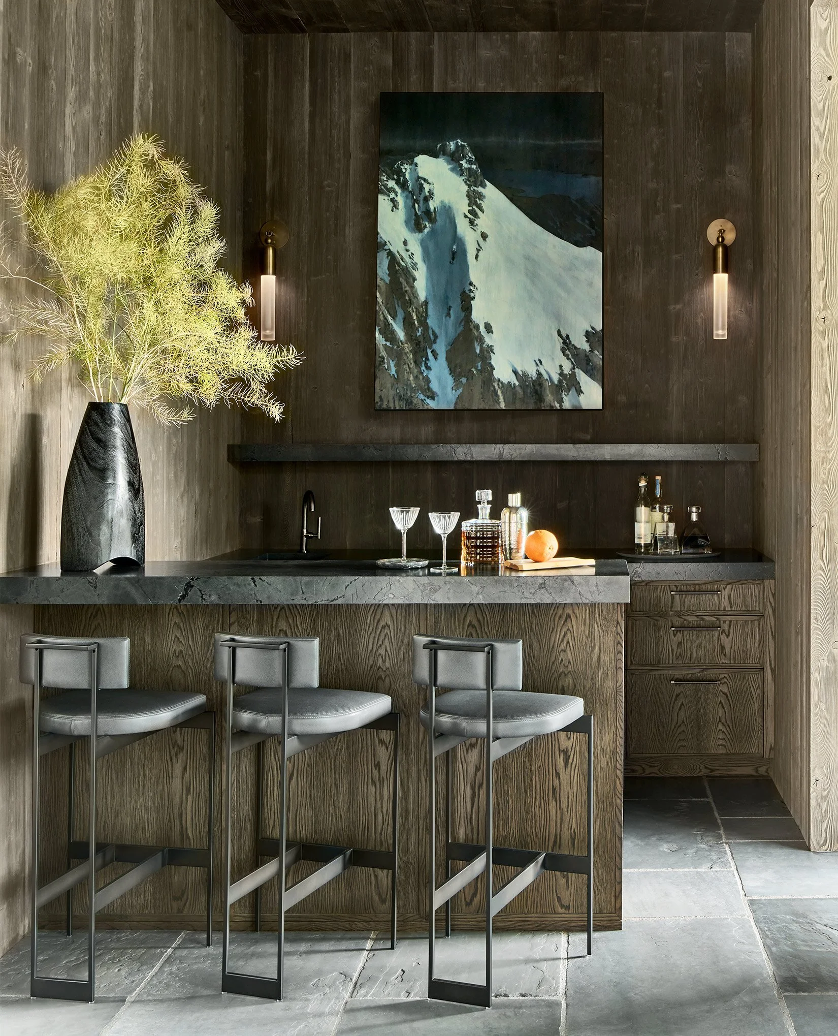 Modern home bar area with dark wooden walls and a black marble countertop. Three gray bar stools are lined up in front. The wall has a large painting of a snowy mountain landscape, flanked by two wall sconces. Decorative items on the counter include 