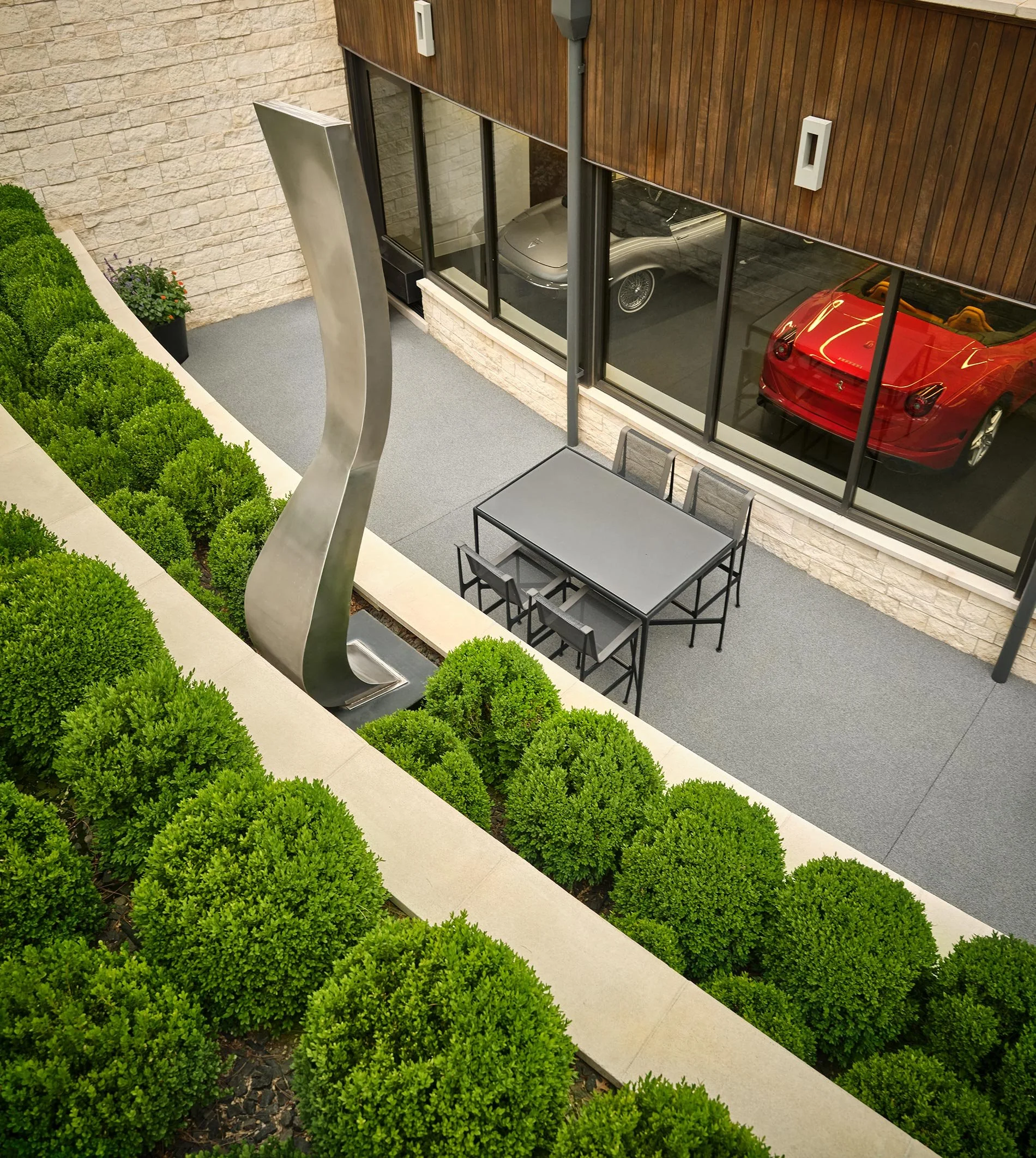 An outdoor patio area with a gray table and chairs, a modern metal sculpture, and well-maintained green bushes, with a view of a garage containing two cars, one red and one silver, through large glass windows.
