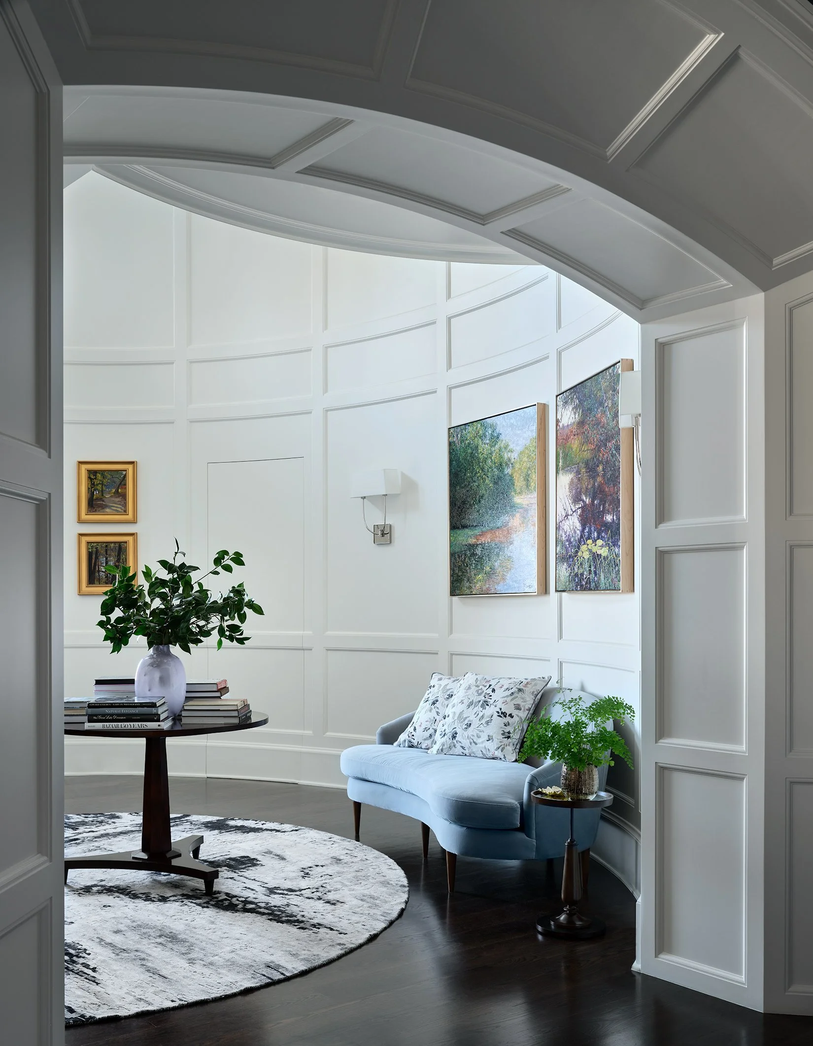 Bright living room with white paneled walls, a light blue sofa with floral pillows, a round rug, a table with books and a potted plant, and artwork on the walls.