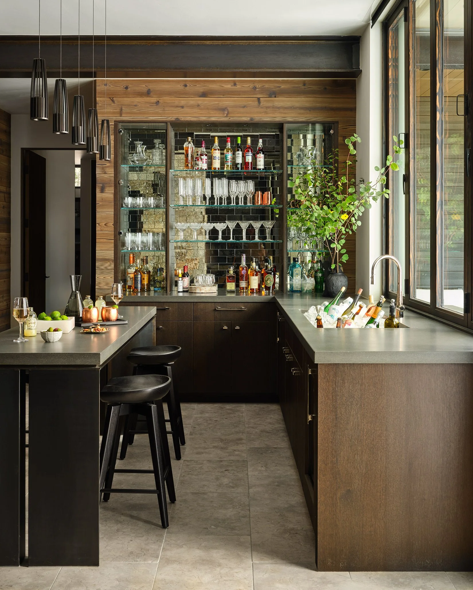 Modern kitchen with dark wood cabinets, a gray countertop, and a large window. Contains a bar area with liquor bottles, glassware, and a potted plant.