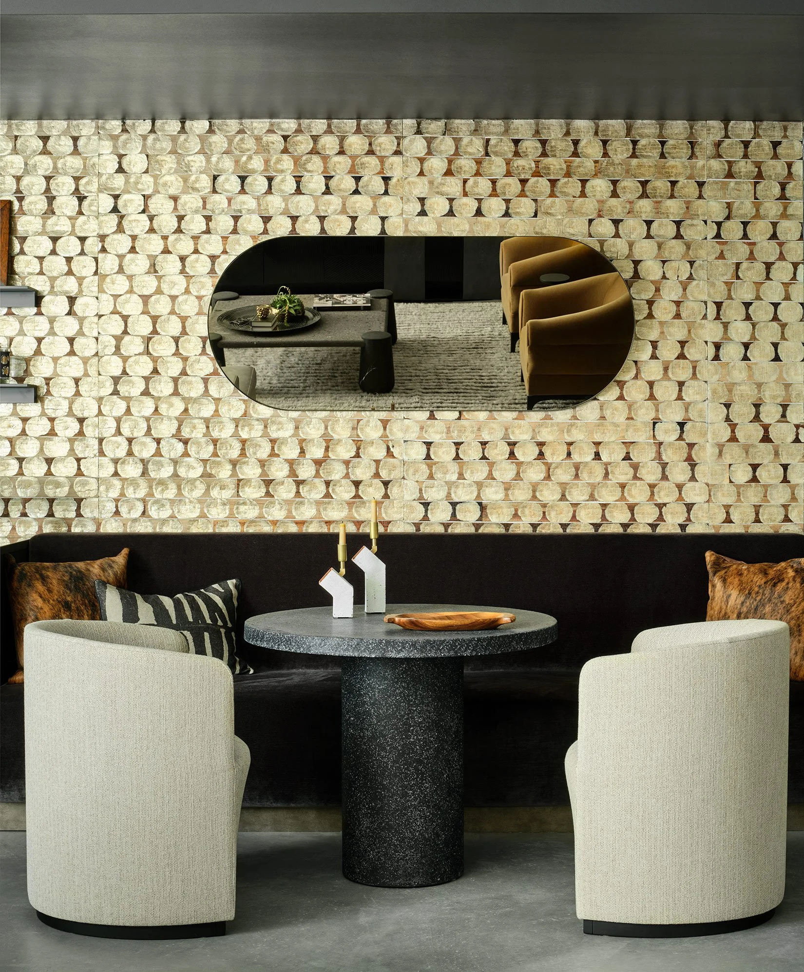 Interior view of a modern dining area with a round black speckled table, two cream upholstered chairs, a black sofa with decorative pillows, a wall mirror, and a patterned brick wall backdrop.