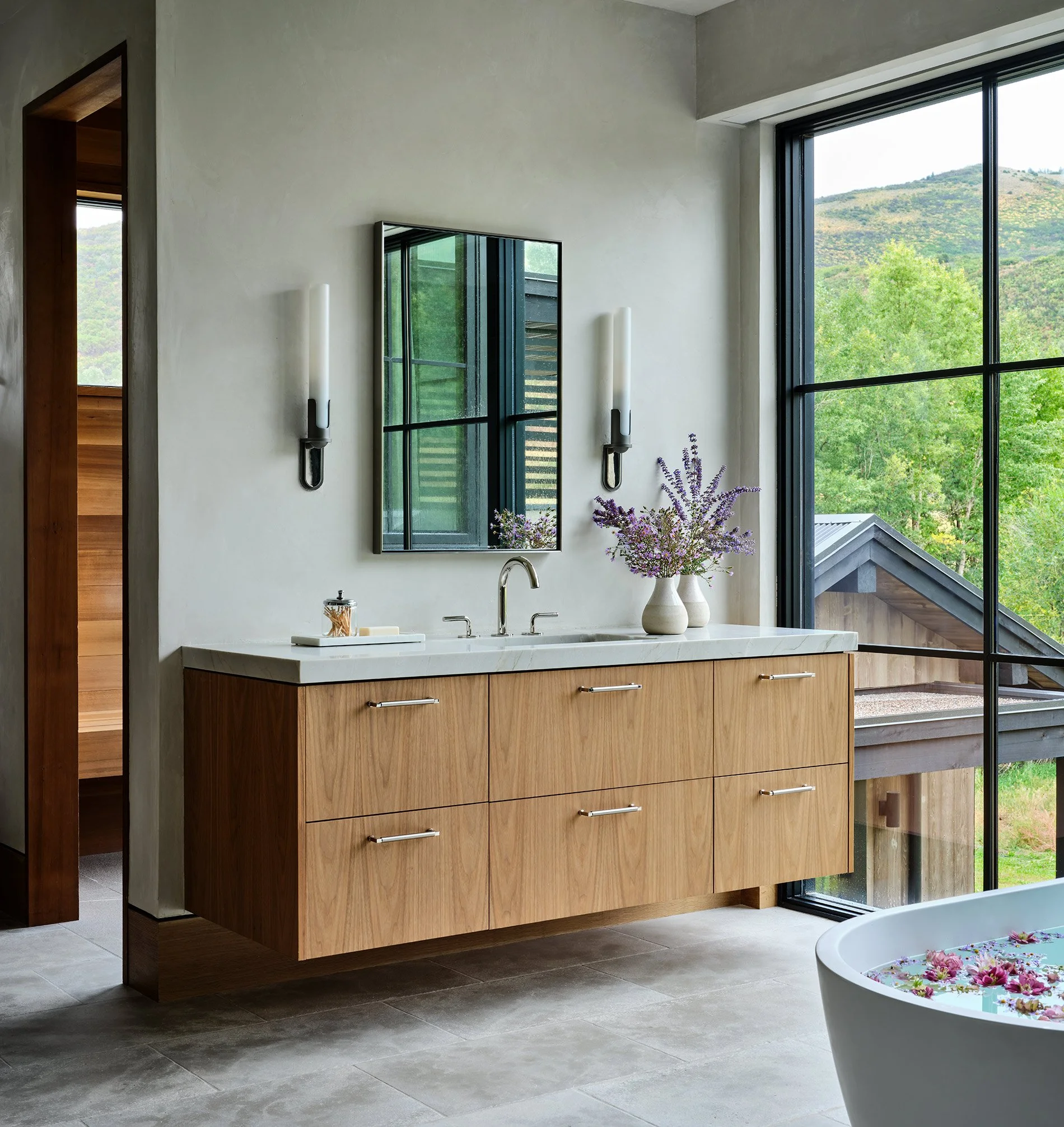 Modern bathroom with a floating wooden vanity, white marble countertop, and a large window showing greenery and a mountain. Decor includes a vase with purple flowers and a large bathtub with floating flowers.