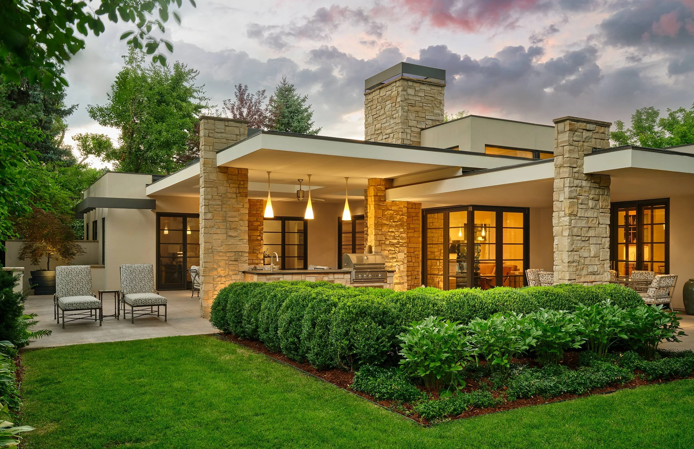 Modern house with stone columns and large glass windows, outdoor patio with chairs and a grill, surrounded by lush green lawn and landscaped bushes, under a partly cloudy sky at dusk.