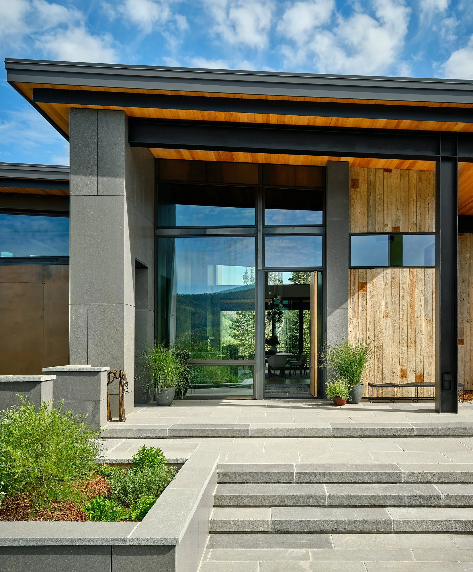 Modern house with large glass windows, wooden accents, and potted plants at the entrance, set against a partly cloudy sky and green landscape.