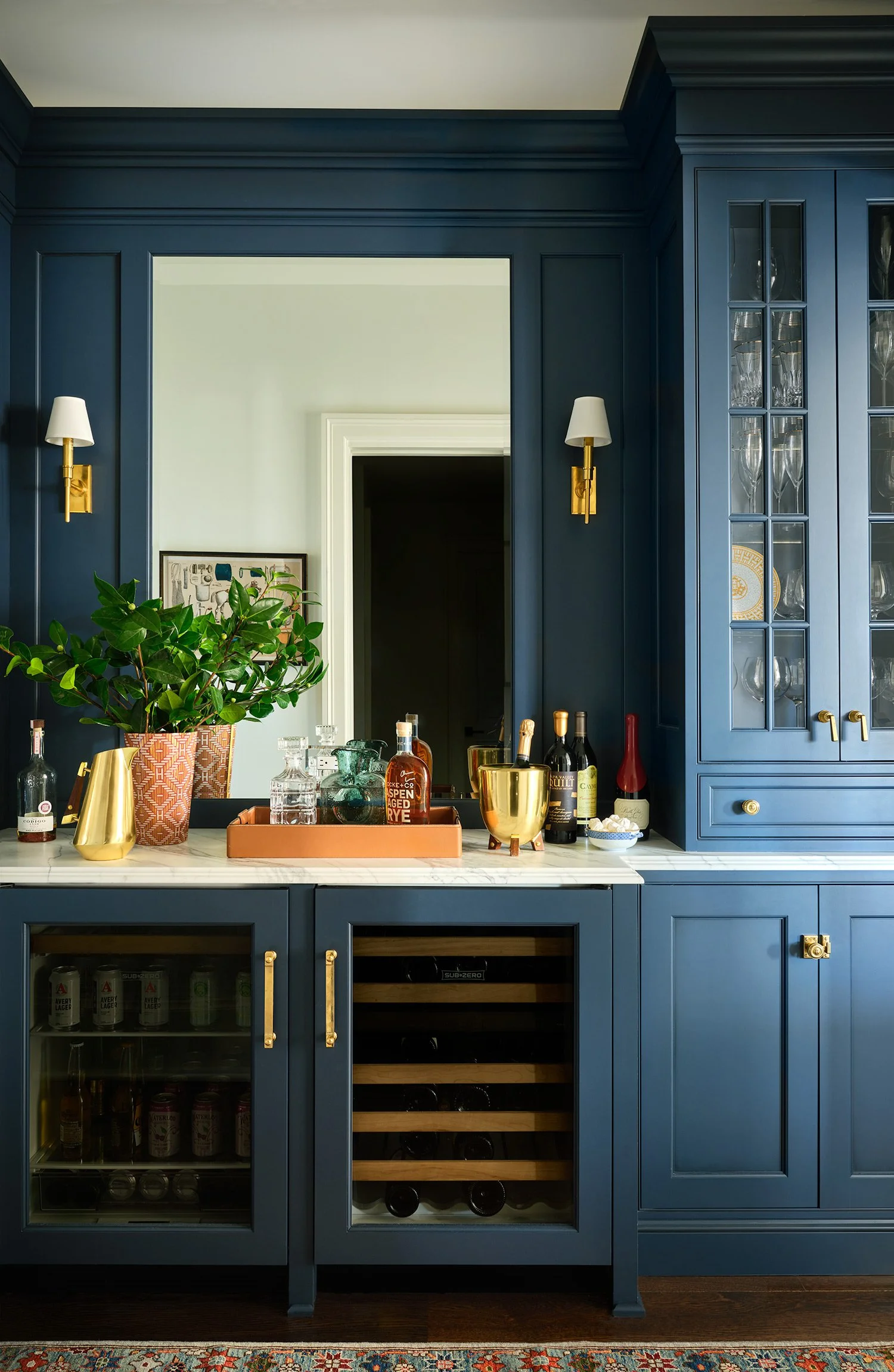 A bar area with blue cabinetry, a white marble countertop, and various bottles and glasses. There are wall sconces with white shades, a large mirror, a potted plant, and a wine fridge.