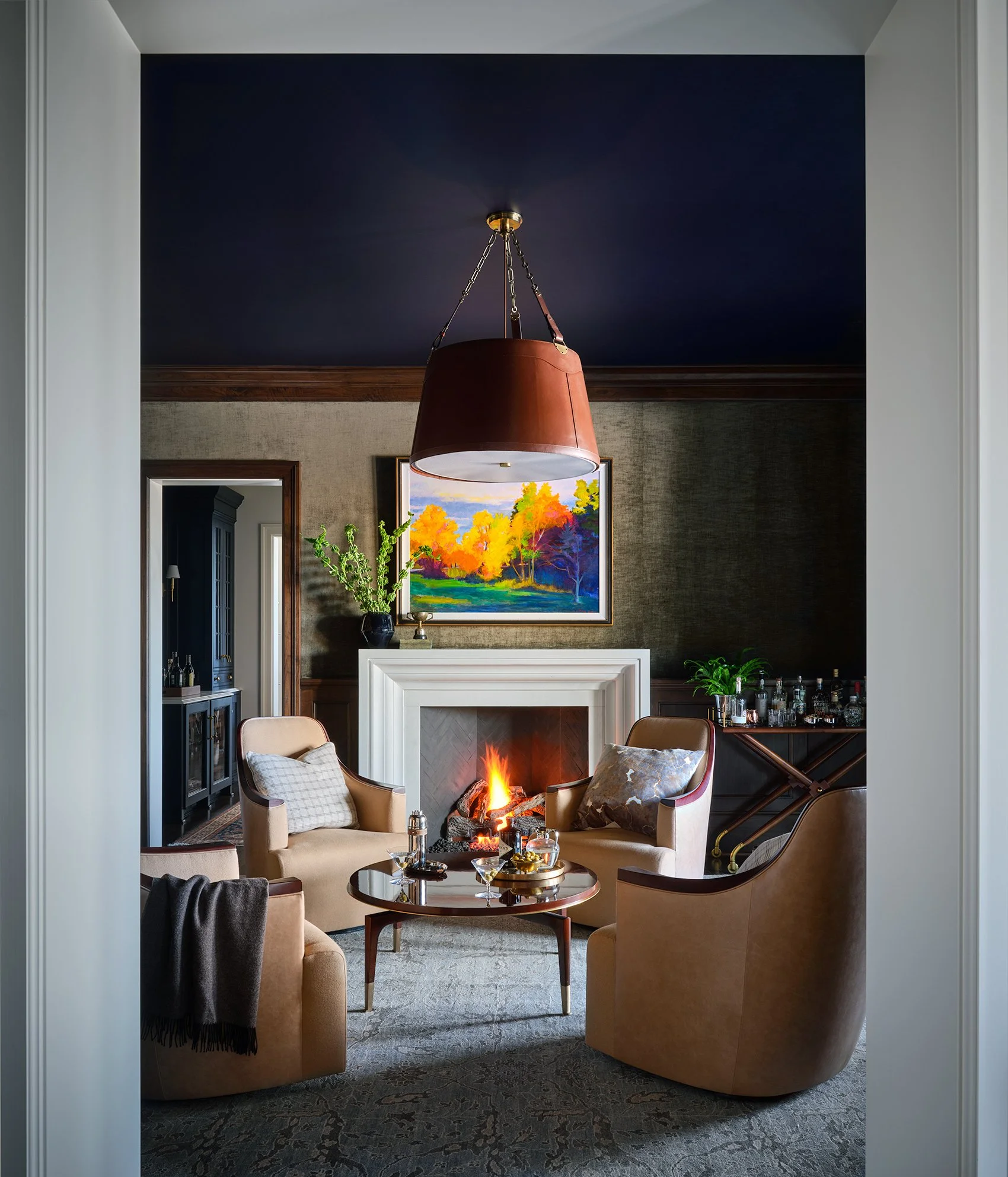 A cozy living room with a fireplace, four armchairs, a coffee table with drinks, a large colorful landscape painting above the fireplace, and a dark ceiling with a hanging lamp.
