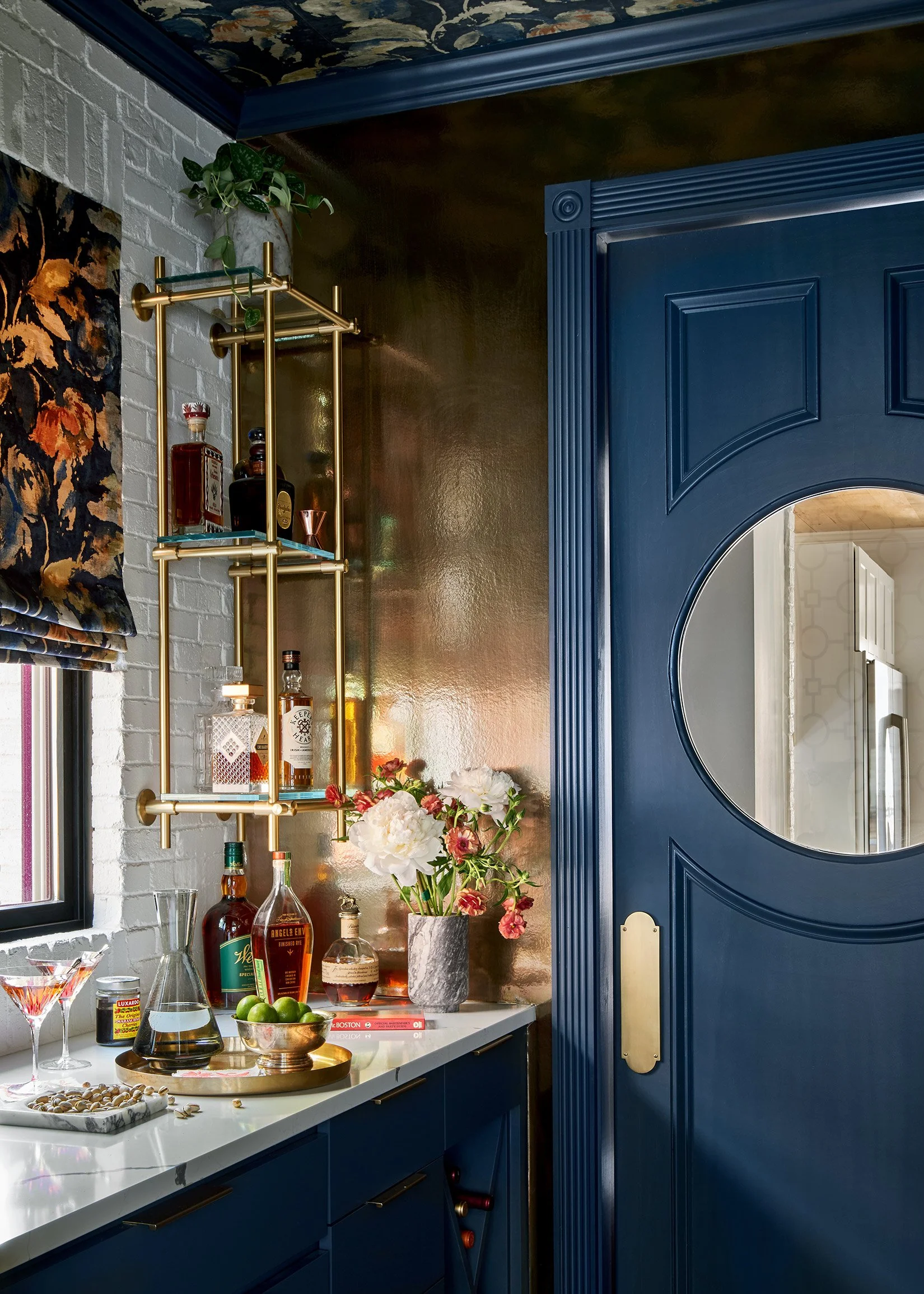 A bar area with a white countertop, navy blue cabinets, and a decorative circular mirror on a blue door. The bar features liquor bottles, a bowl of limes, flowers, and bar tools. A gold shelving unit holds liquor bottles and a plant on top. There's a