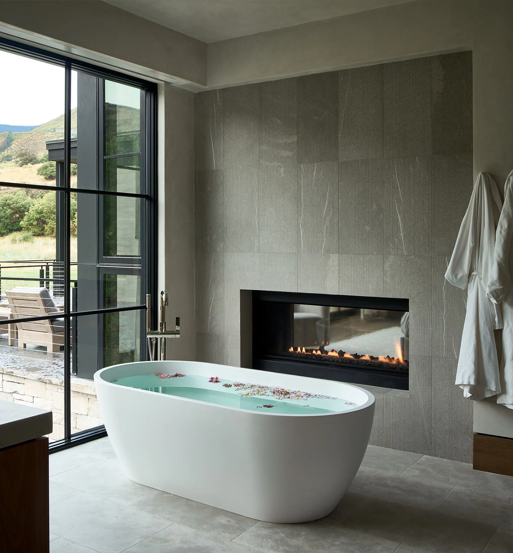 Modern bathroom with a freestanding bathtub filled with water and flower petals, next to a large window overlooking a scenic outdoor landscape, with a fireplace and white bathrobes hanging on the wall.