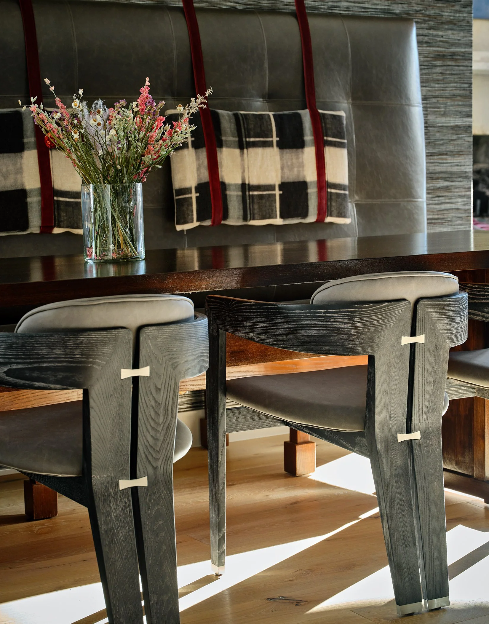 A dining area with a dark wooden table, black chairs with beige cushions, and a vase of mixed flowers on the table. There are pillows with black, white, and red plaid pattern against a padded, dark leather wall.