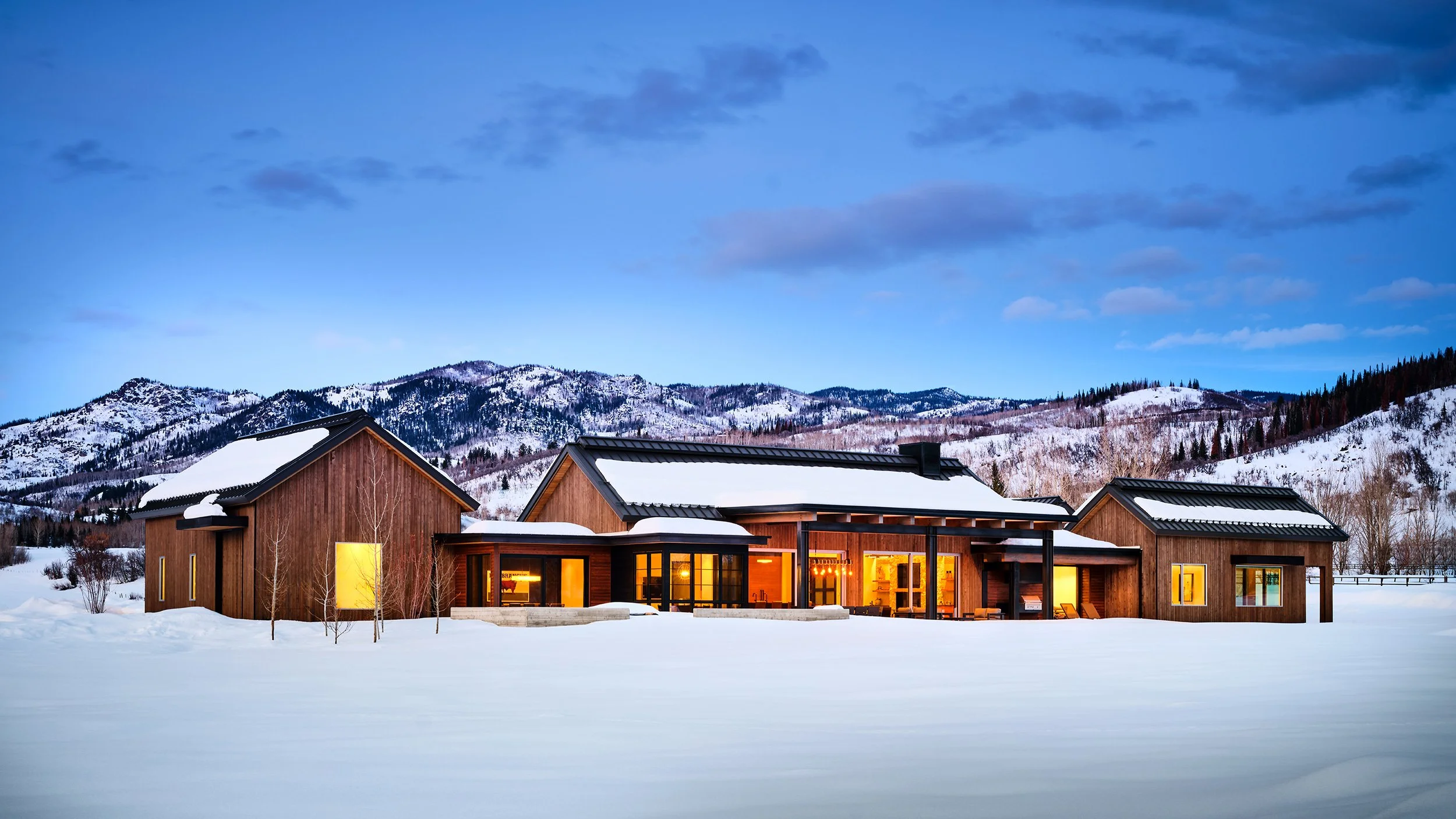 Modern wooden house with large windows in a snowy landscape, mountains in the background, and warm indoor lighting visible through the windows.