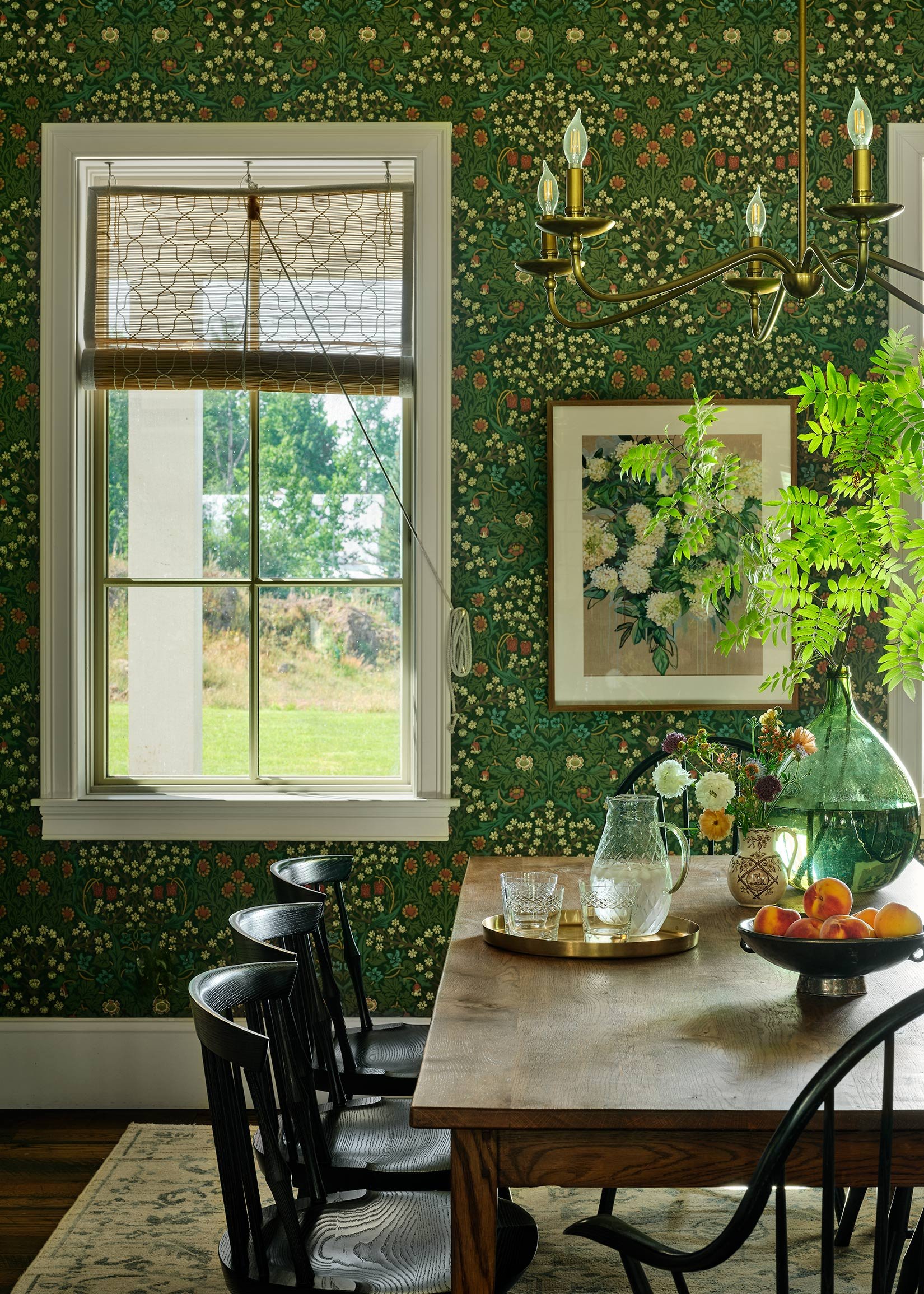 Interior of a dining room with a wooden table, black chairs, green floral wallpaper, a framed floral artwork, a chandelier, a window with a bamboo blind, and decorative vases with flowers and a bowl of peaches.