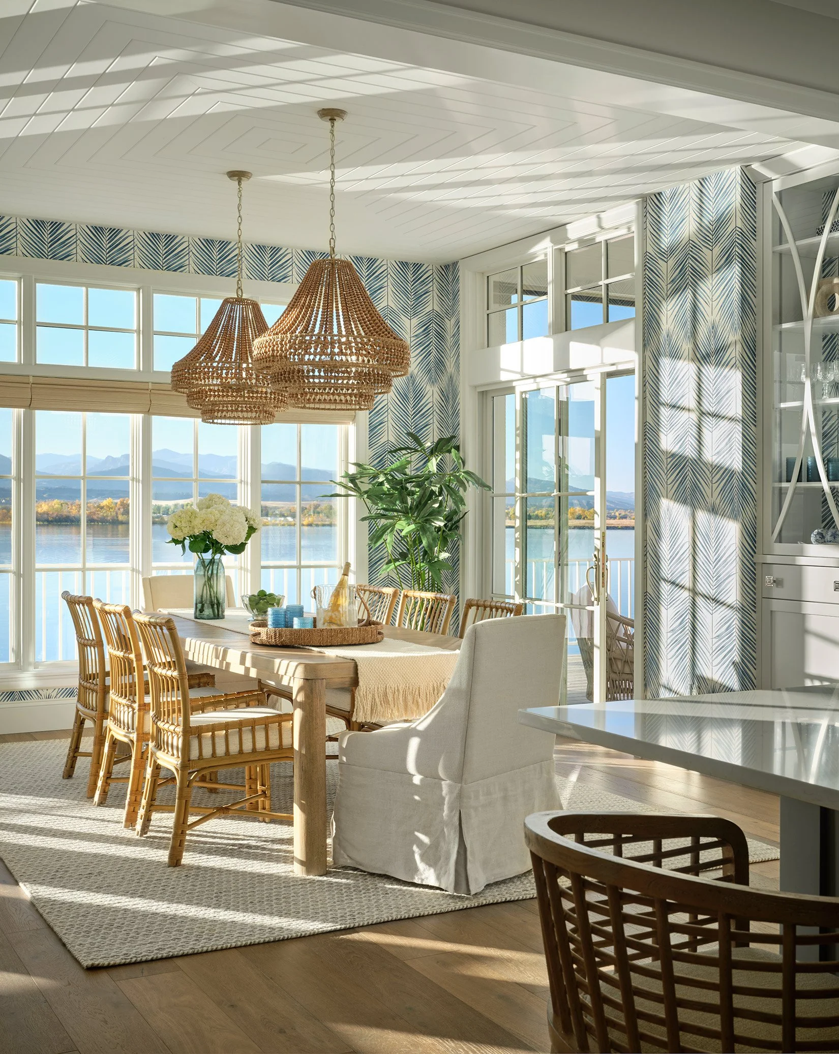 Bright dining room with large windows overlooking water and mountains, featuring a wooden dining table with wicker chairs, hanging wicker pendant lights, a glass vase with white flowers, and patterned wallpaper.