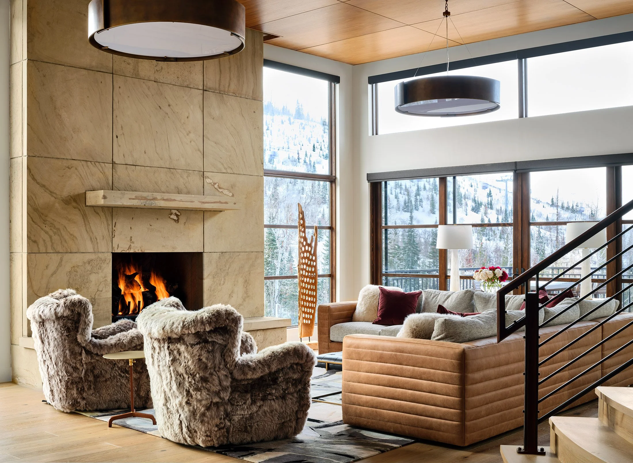 A cozy living room with a large stone fireplace, two plush furry armchairs, a beige sofa with throw pillows, and large windows showing snowy mountains outside.