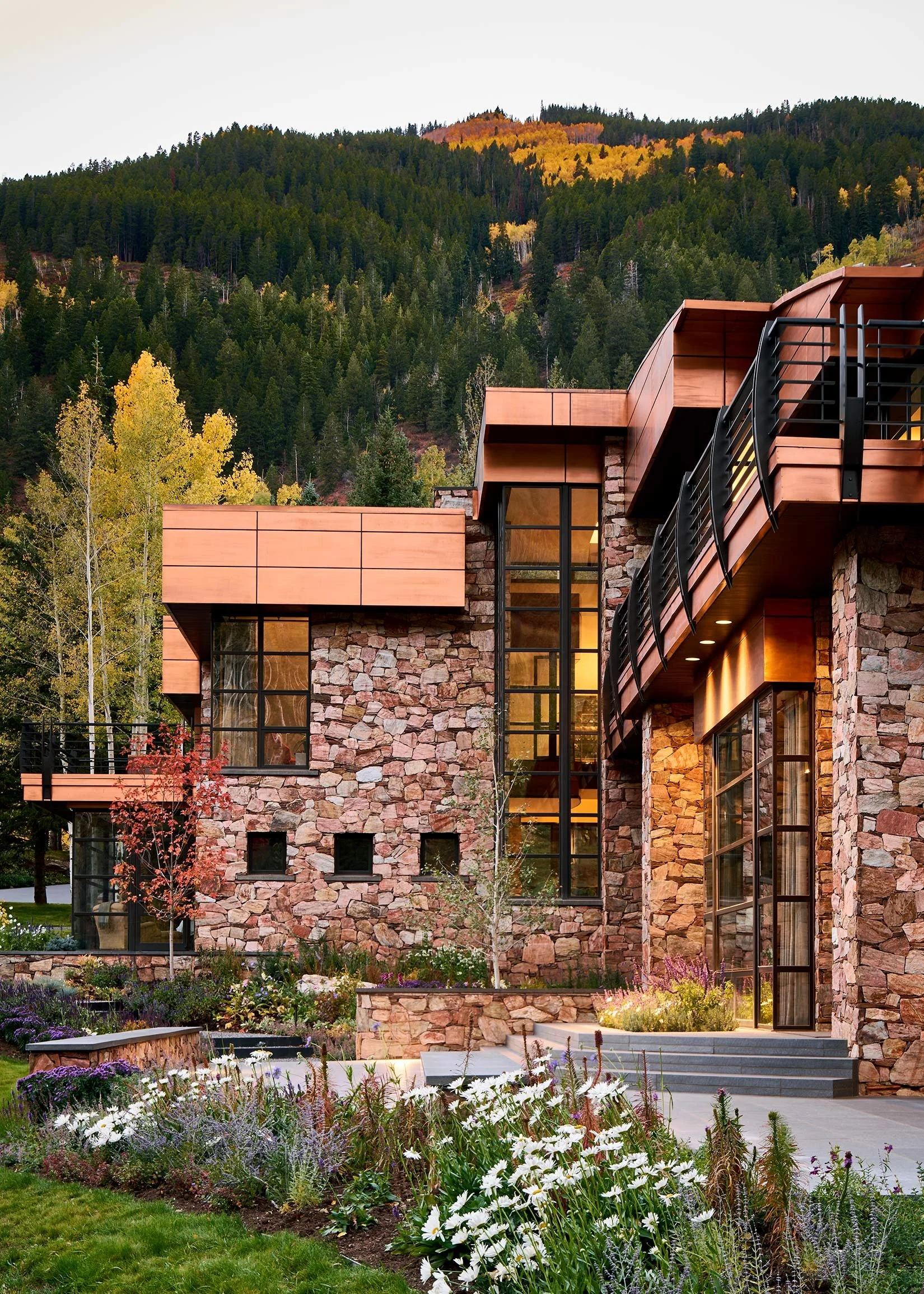A modern house with stone and wood exterior, large windows, and a balcony, set against a mountain with trees and colorful autumn foliage, garden with flowers in the foreground.