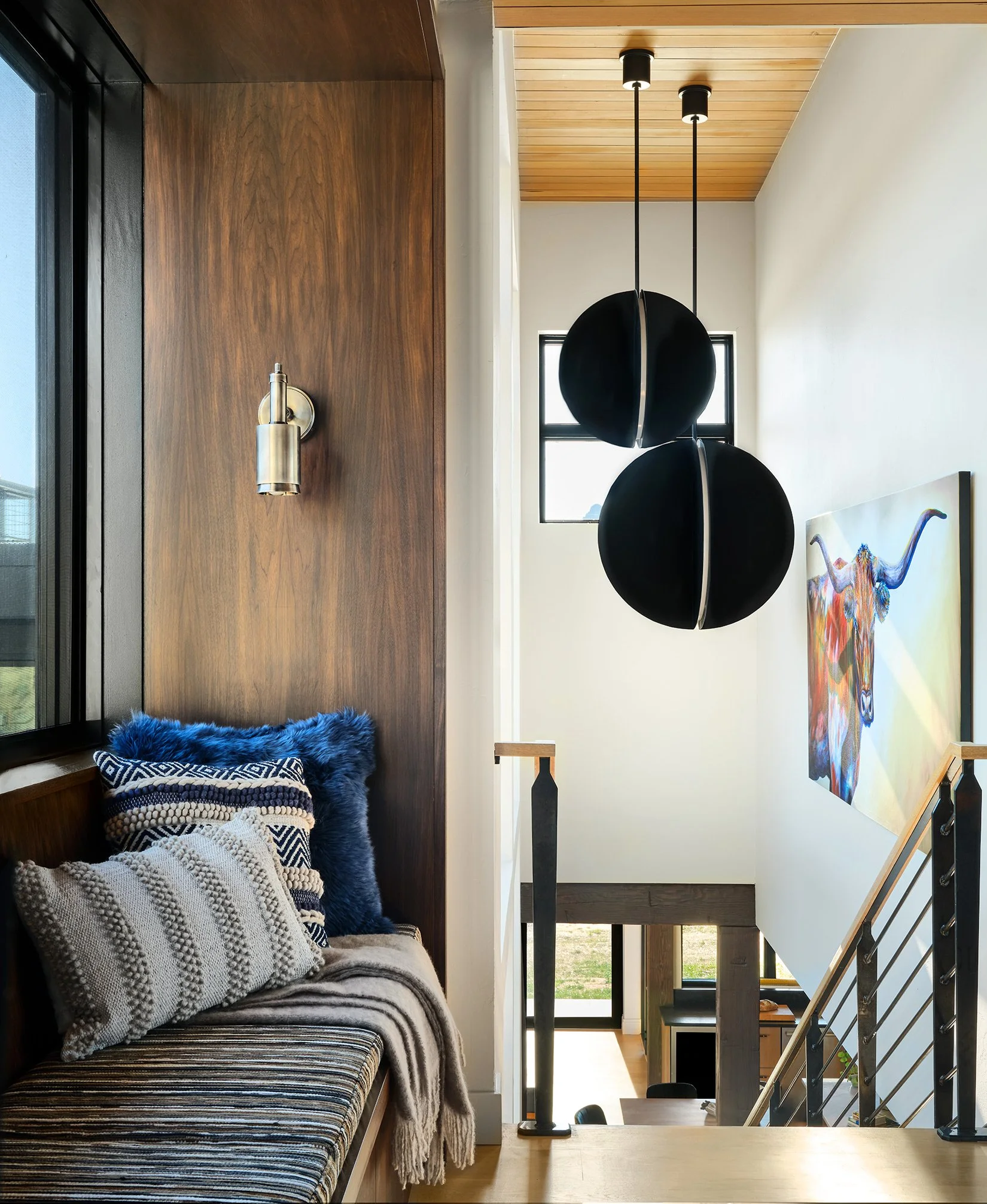 Cozy interior staircase landing with a bench, decorative pillows, a wooden wall, hanging black spherical pendant lights, and artwork of a colorful cow on the white wall.