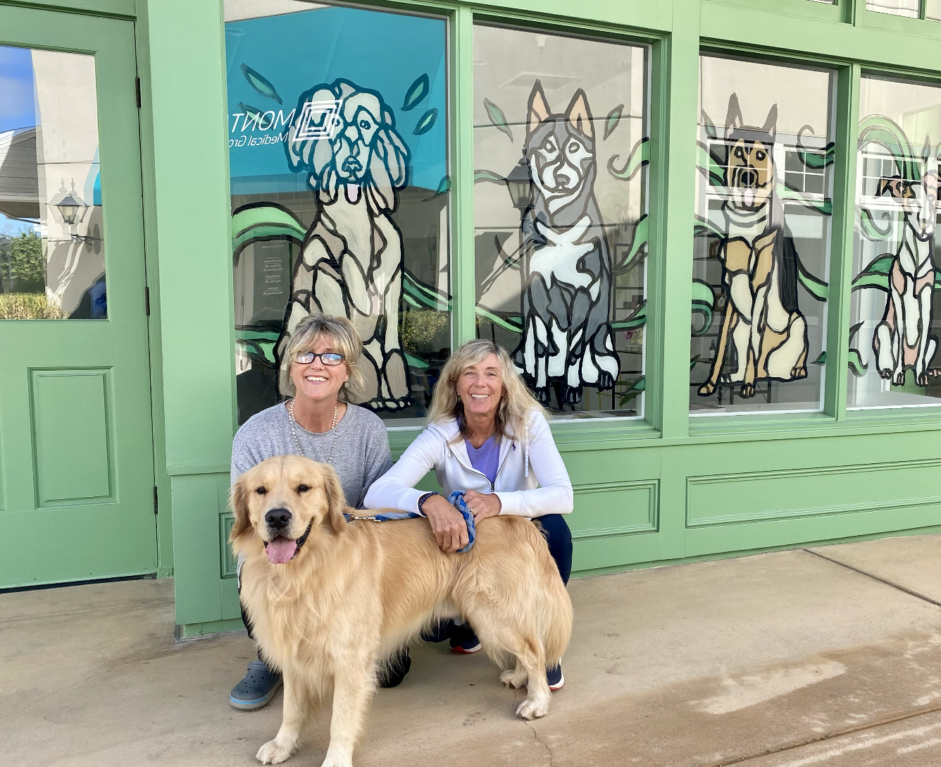 Two smiling women with a golden retriever dog in front of a green storefront with large windows featuring colorful dog illustrations.