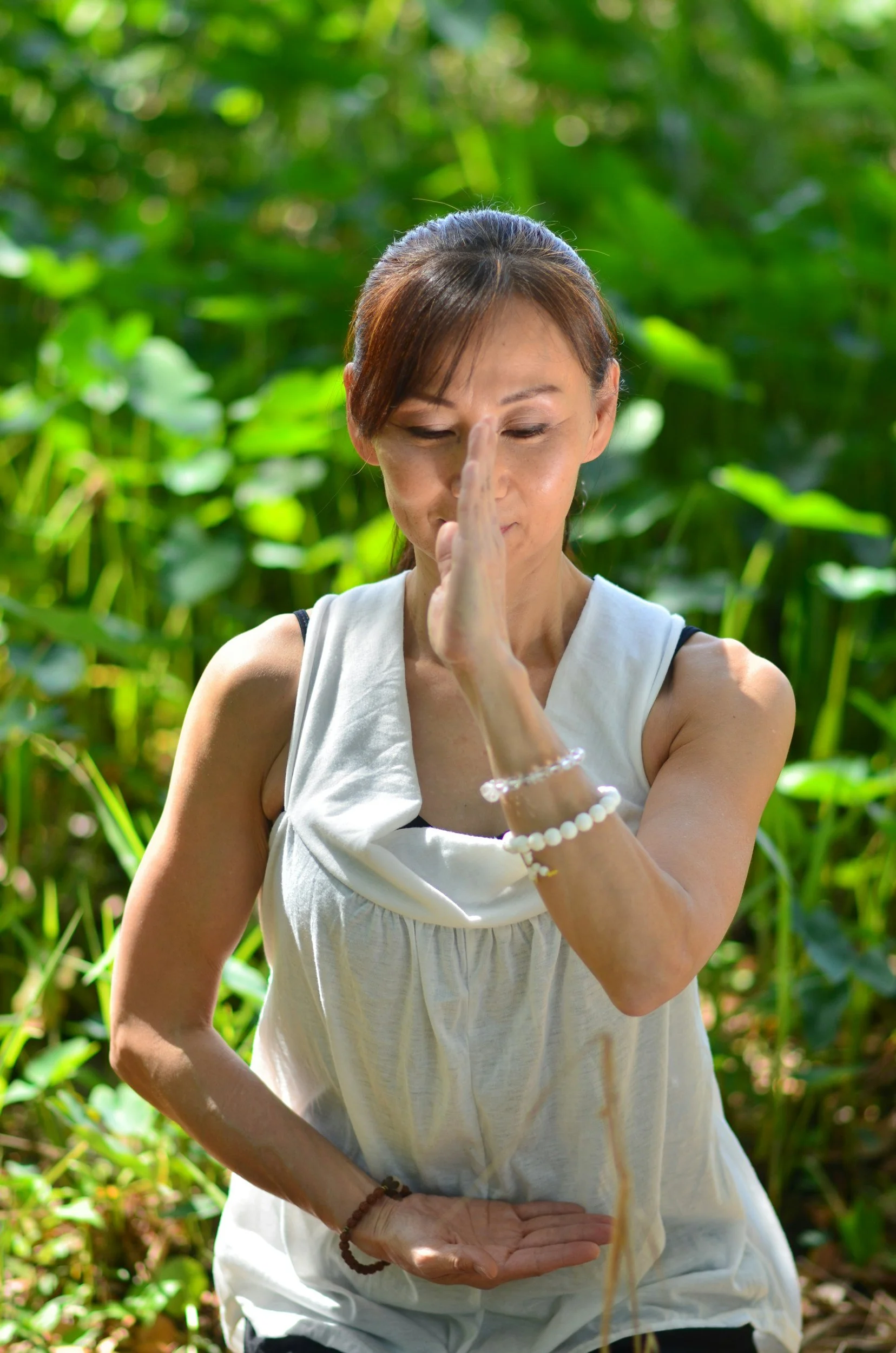 Woman practicing yoga outdoors in a lush green setting, standing with her eyes closed, hands pressed together in a prayer pose.