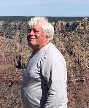 An older man with white hair standing near the Grand Canyon, smiling and wearing a light gray long-sleeve shirt.